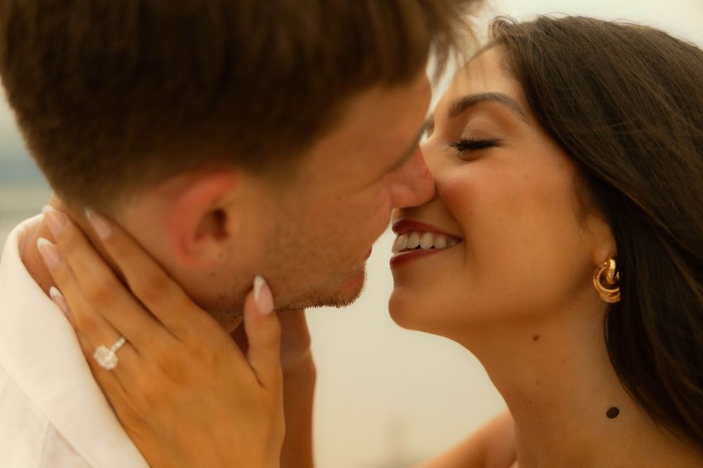 Couple sharing a romantic moment, woman smiling with engagement ring visible, close face-to-face pose.