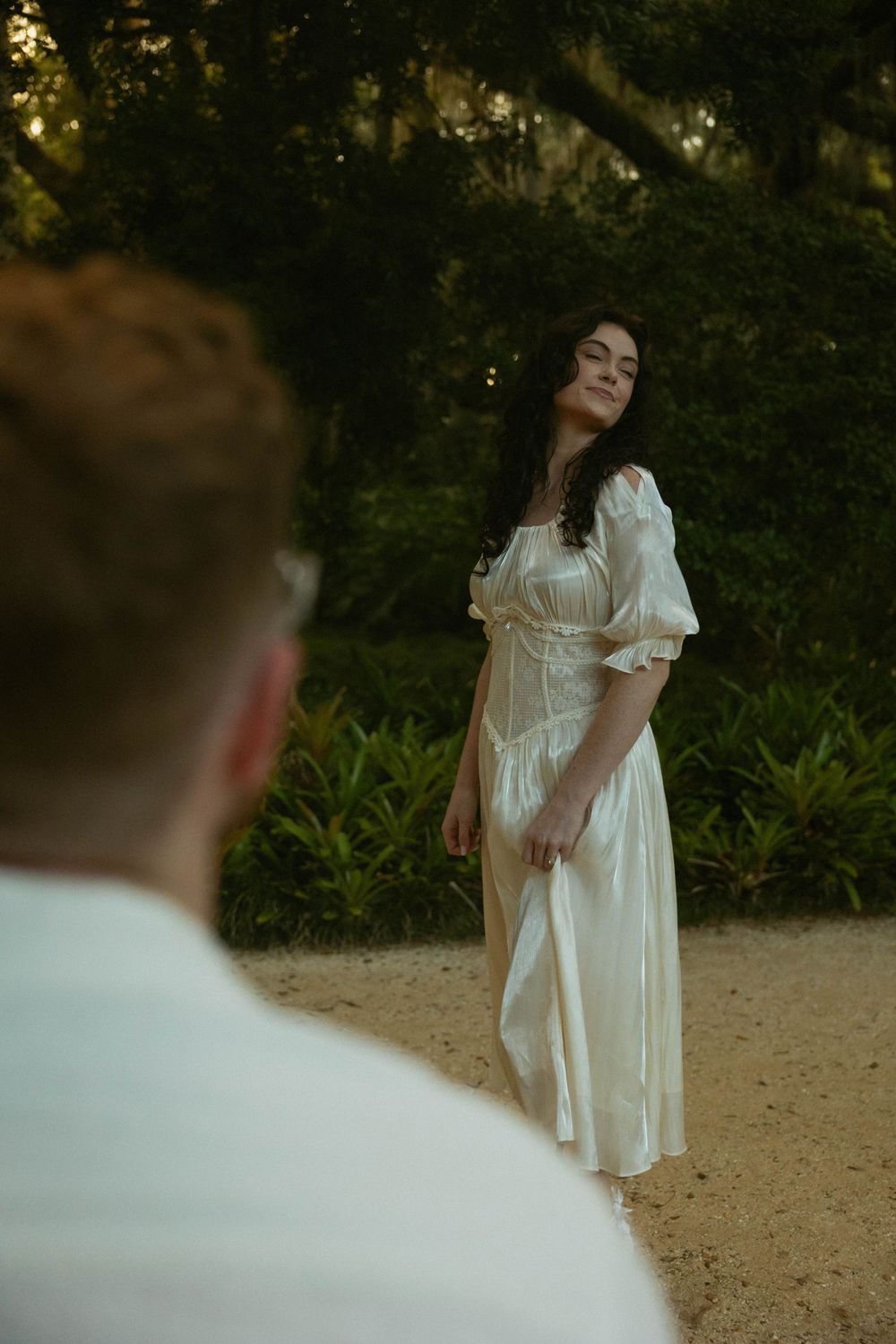 Woman in white satin dress stands gracefully on sandy path surrounded by lush greenery, with blurred figure in foreground.