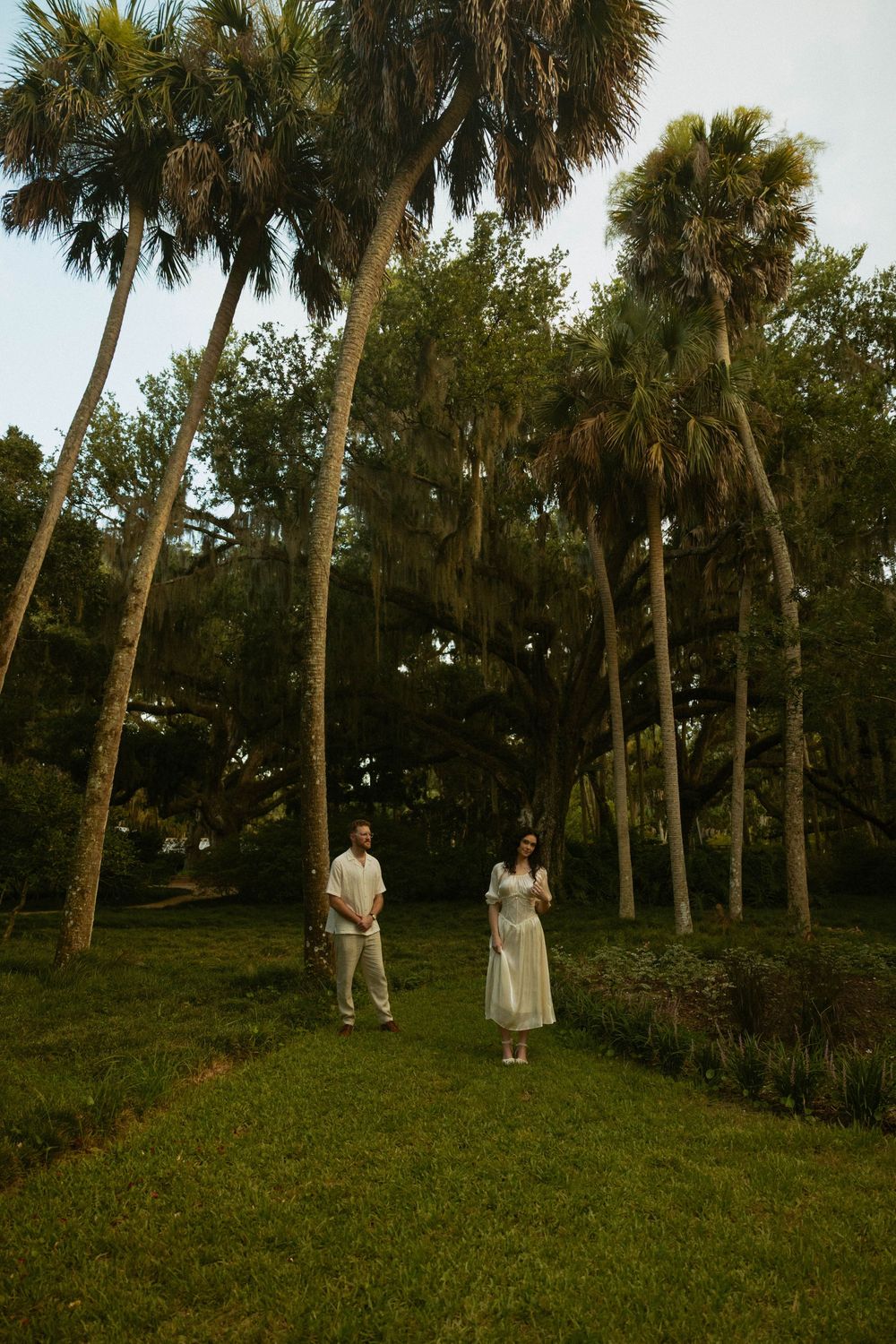 A couple in white attire stands on a lush green lawn surrounded by tall palm trees in a serene garden setting.