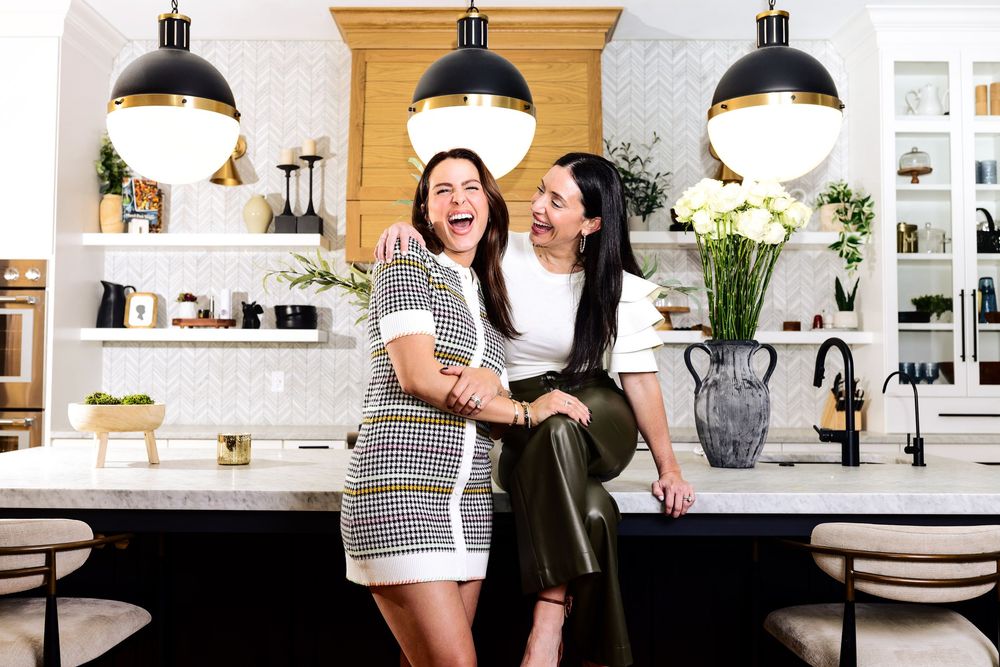 Two friends laughing together in a modern white kitchen with pendant lights.