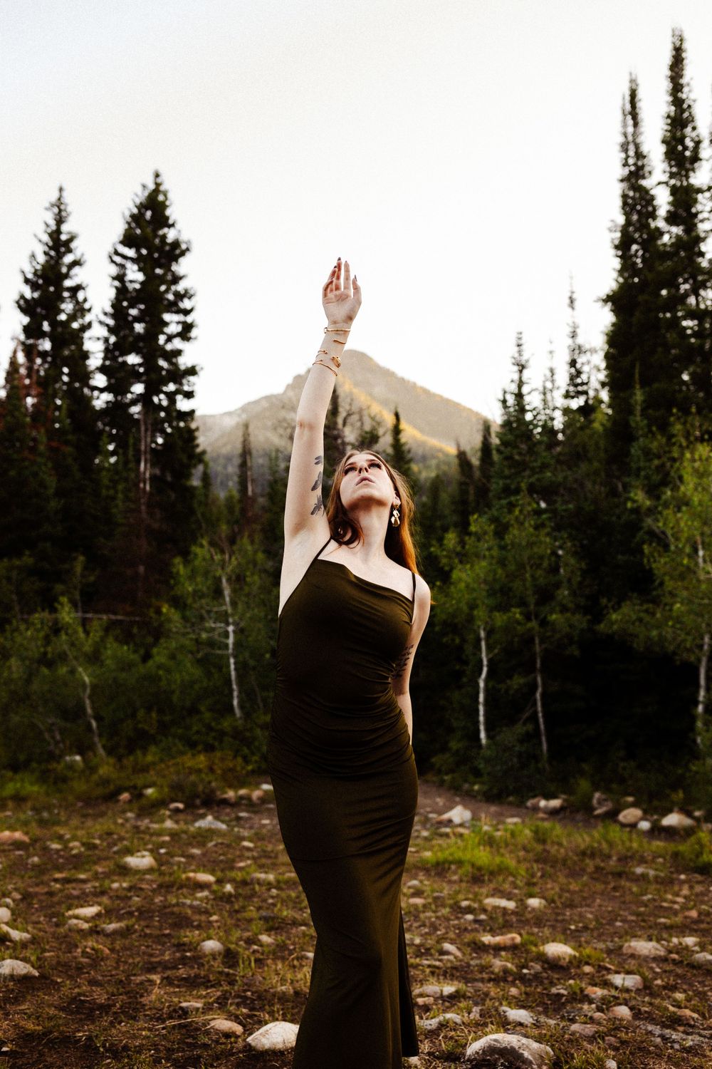 Dramatic pose in a black dress against a mountain landscape with evergreen trees.