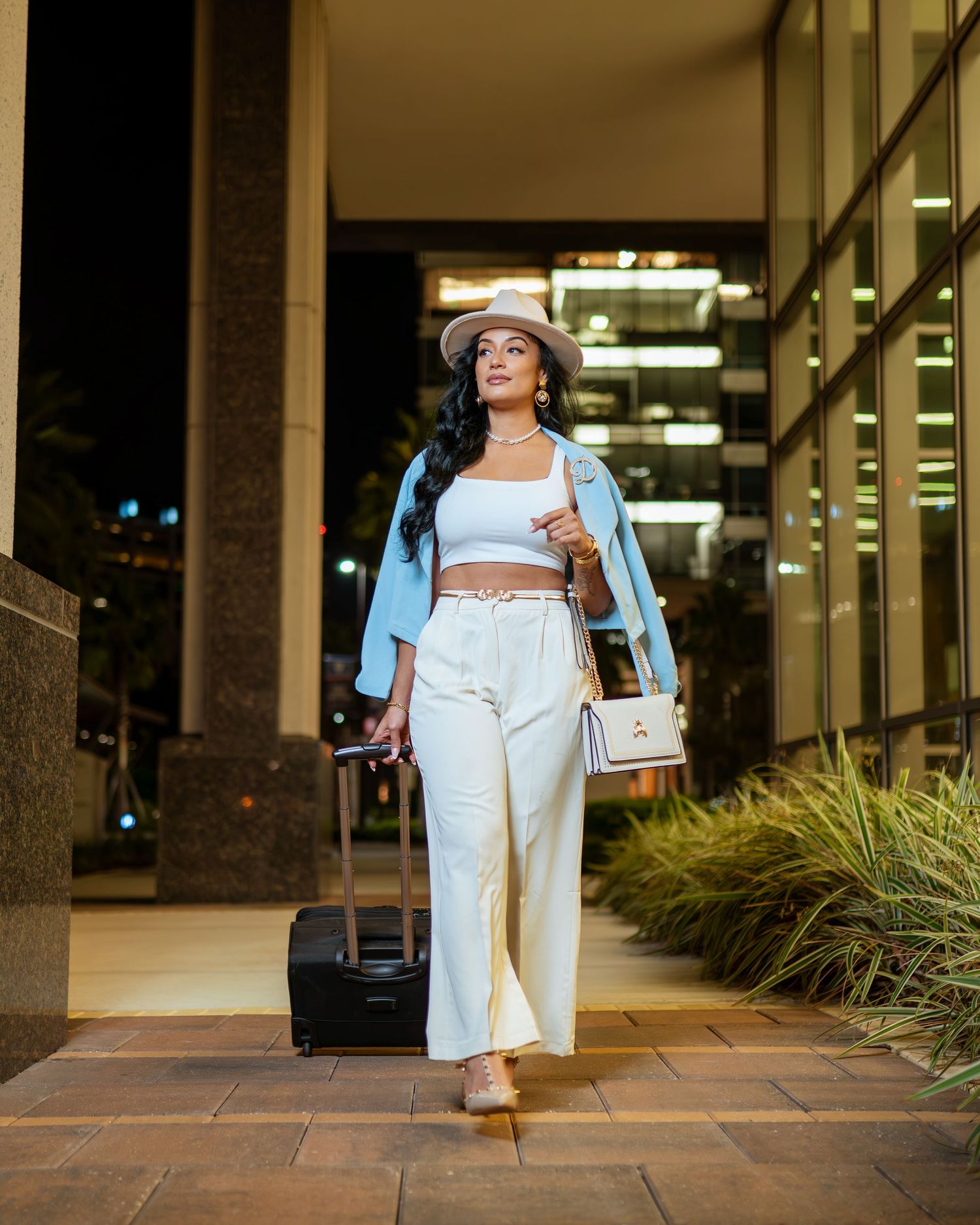 Stylish traveler in light blue jacket and white pants walks confidently through outdoor courtyard with rolling luggage.