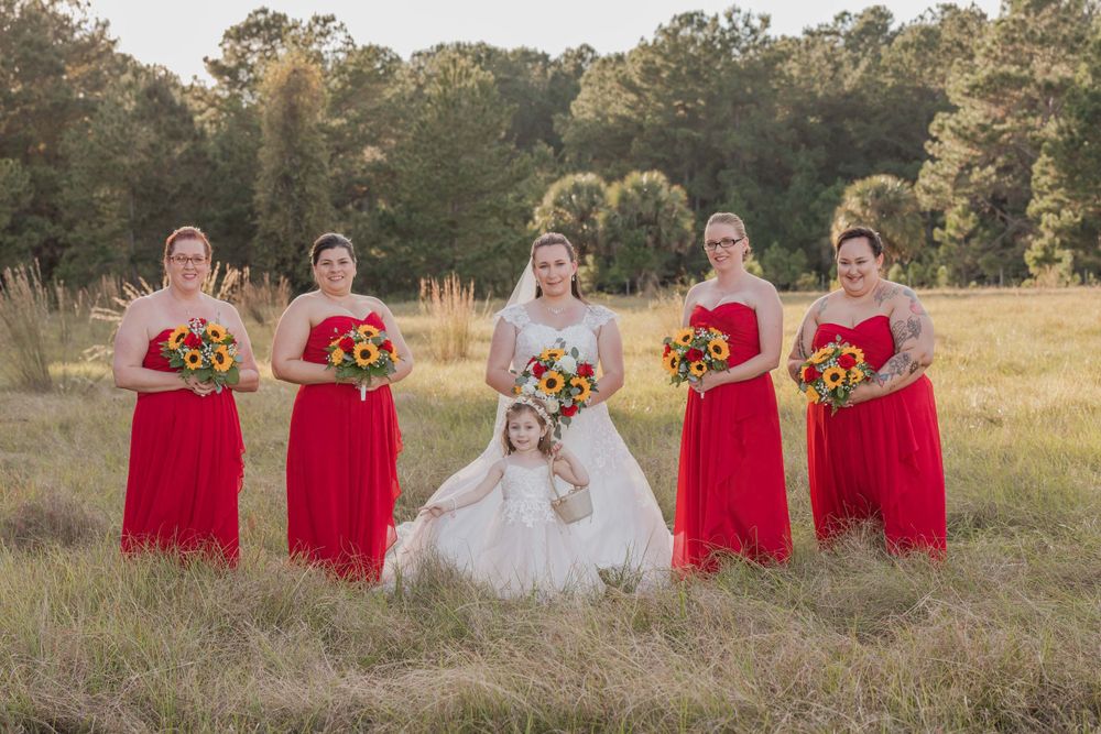 A wedding party stands in a sunlit field with bridesmaids in red gowns holding yellow sunflower bouquets.