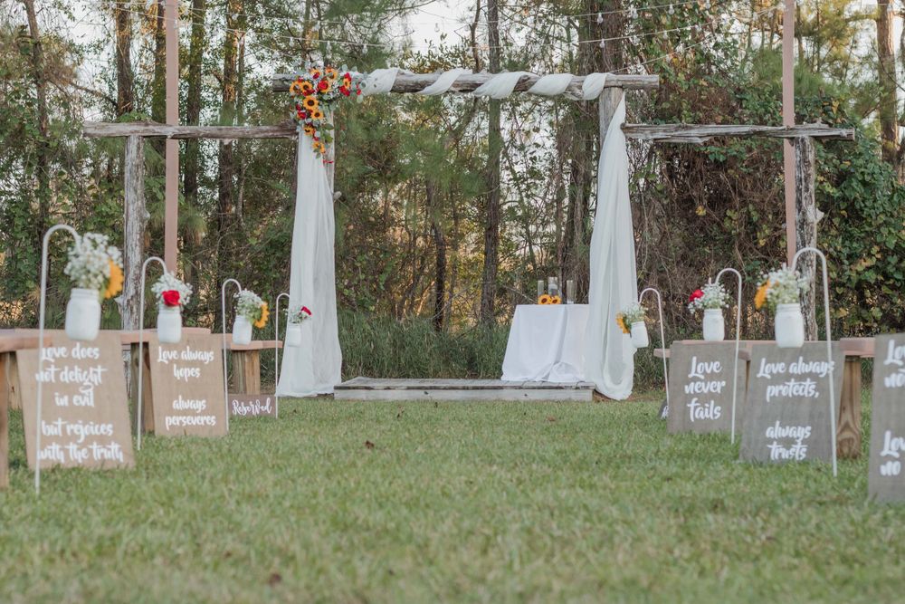 Rustic outdoor wedding ceremony setup with wooden signs and white fabric draped archway in a natural forest setting.