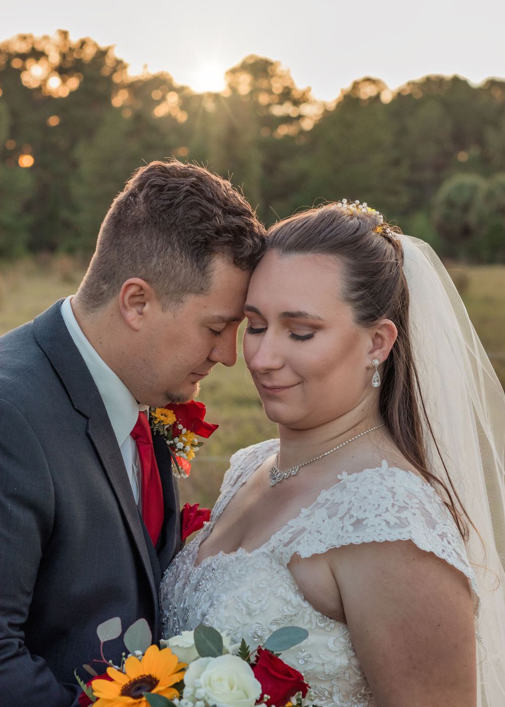 A romantic sunset wedding photo featuring a couple in formal attire with sunflowers and red roses in their bouquet.