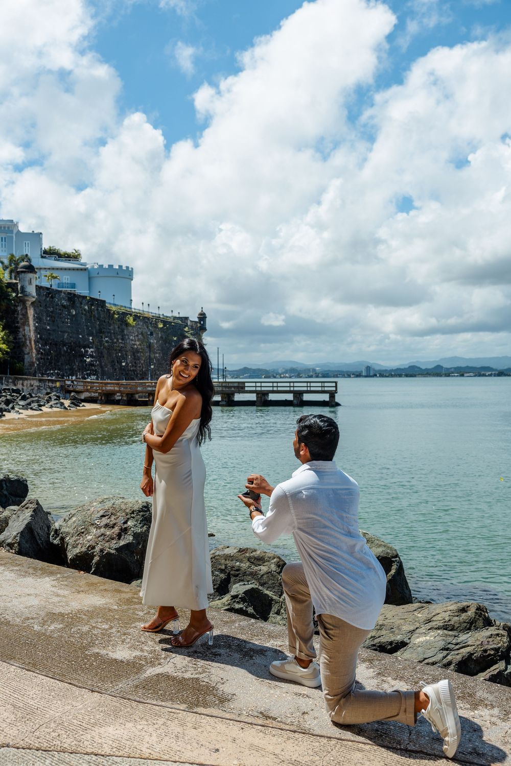 Man proposing on one knee to woman in white dress beside turquoise ocean waters.