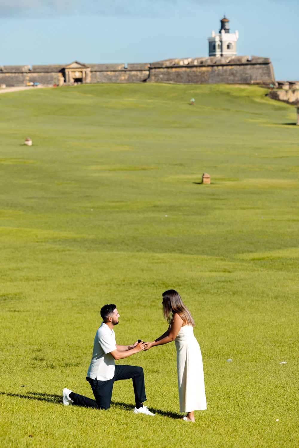 Man proposing on one knee to woman on green lawn near historic fort in Puerto Rico.