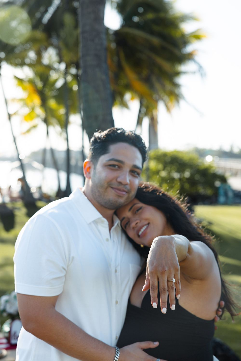 Happy engaged couple embracing under palm trees with woman displaying diamond engagement ring.