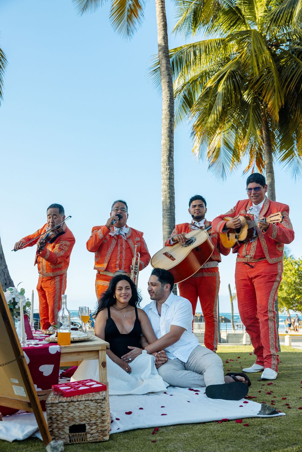 Couple sitting together on blanket while mariachi band in red suits performs under palm trees.