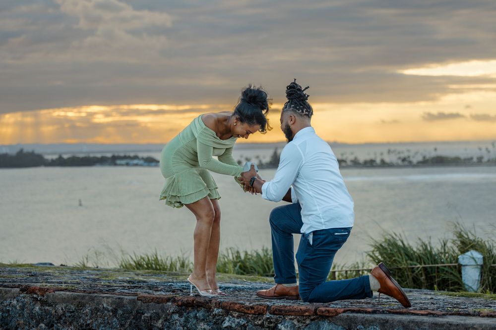 Man proposing on one knee to woman on rocky shoreline during dramatic golden sunset.