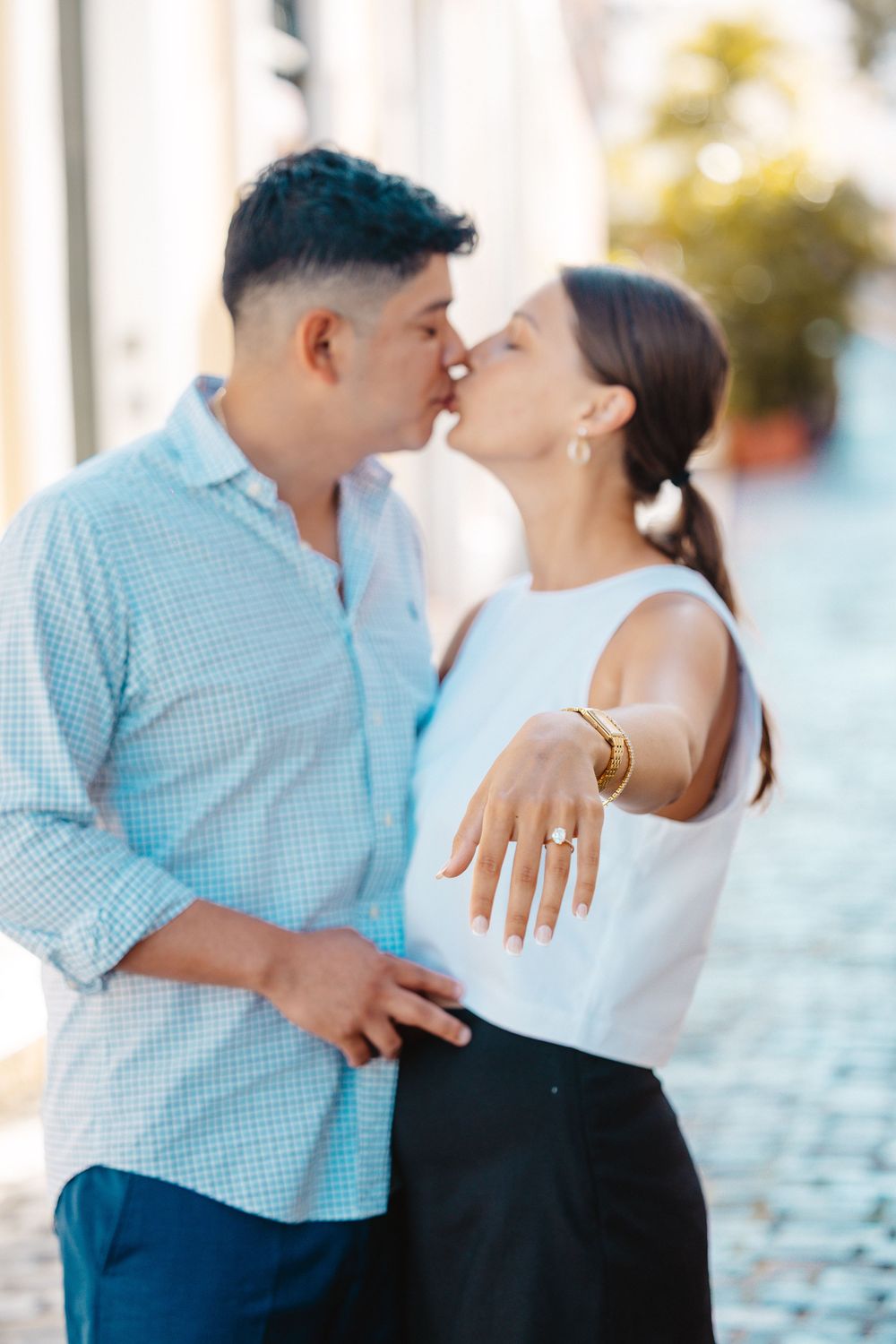 Engaged couple kissing while woman shows off diamond engagement ring on cobblestone street.