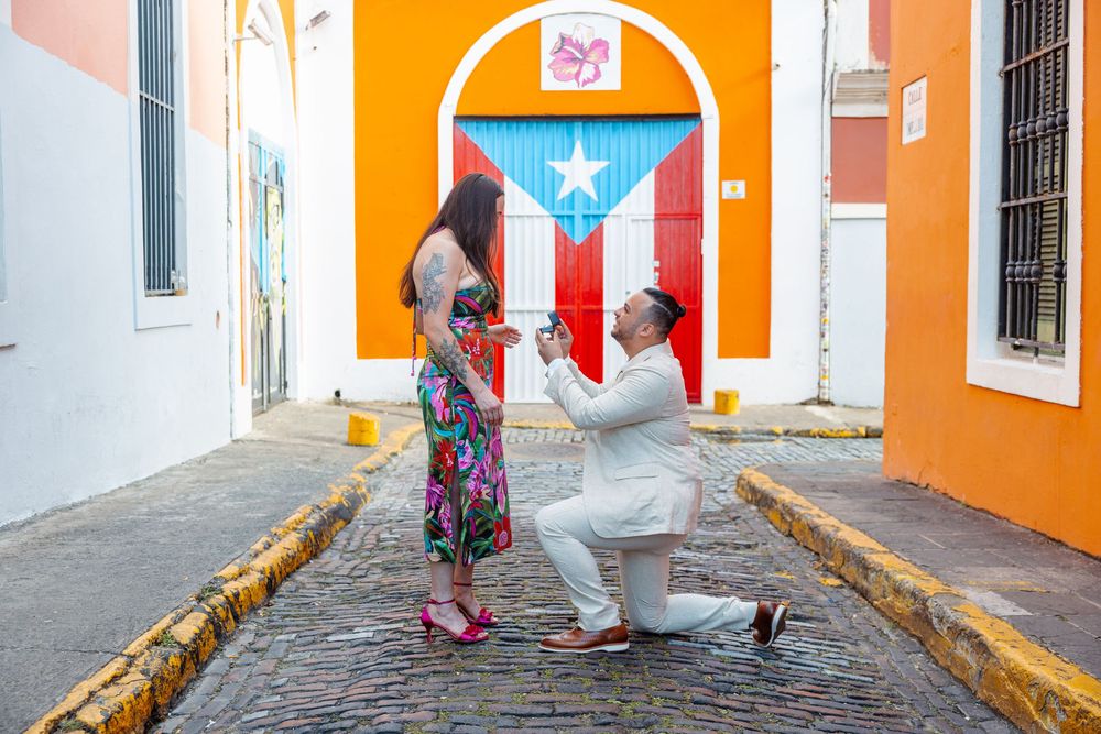 Man proposing on one knee on cobblestone street with colorful Puerto Rican flag mural backdrop.