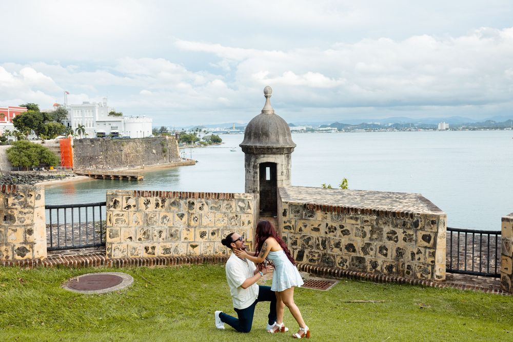 A man proposes to his partner on the grass near a historic stone fortress overlooking the bay.