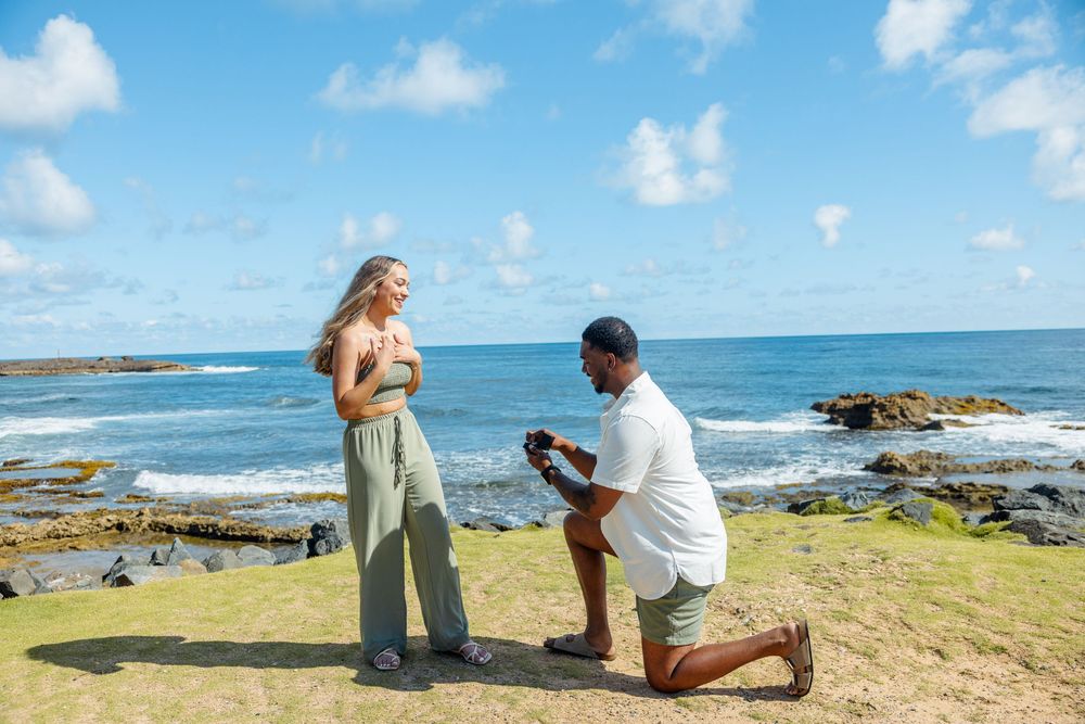 A man proposes on one knee to a woman on a scenic rocky coastline under a bright blue sky.