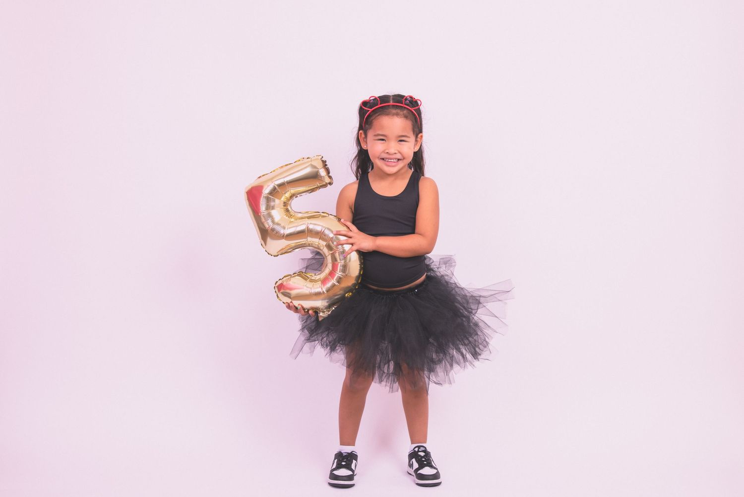 A child in a black tutu dress holds a gold number 5 balloon against a pink background.