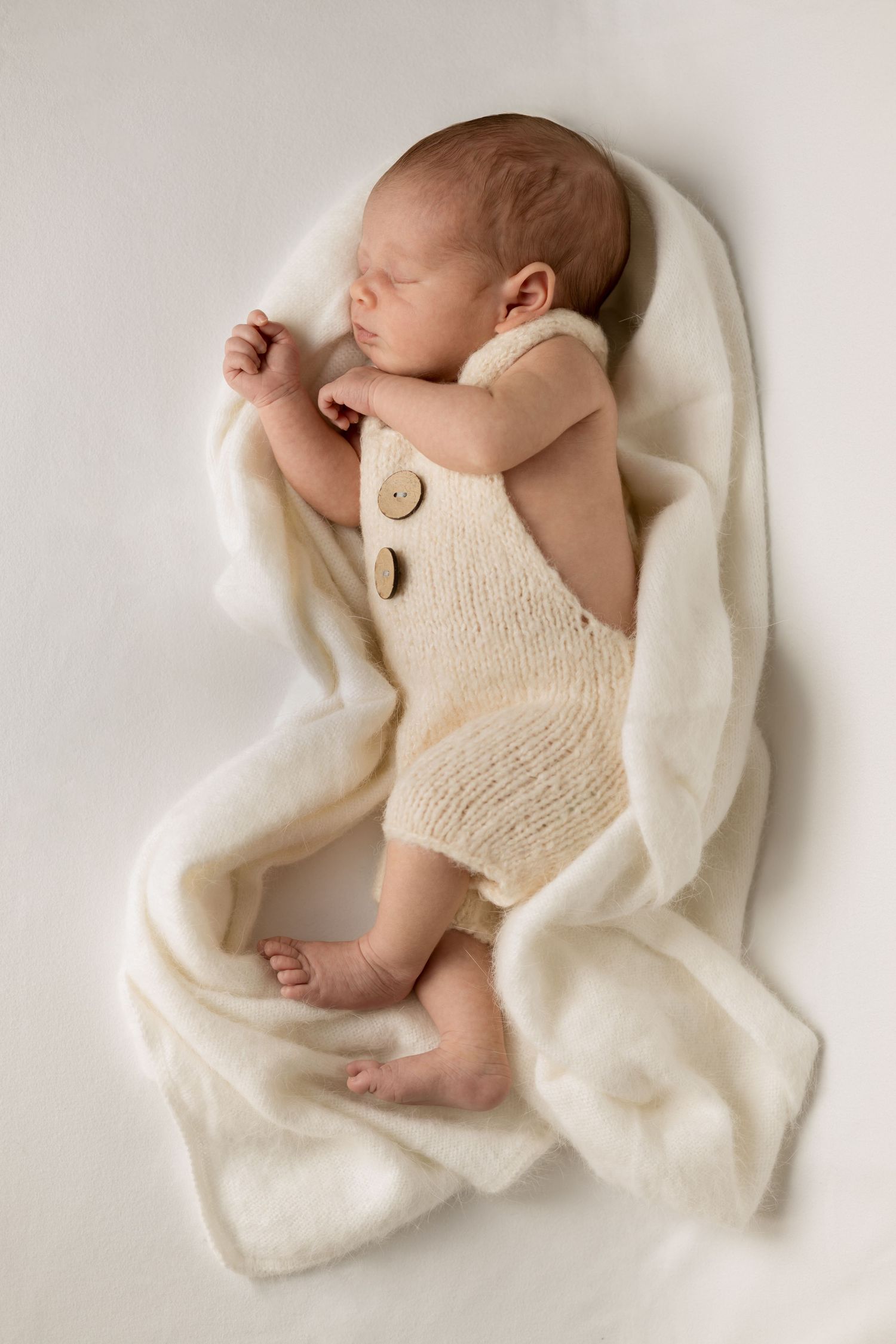 Newborn photography of a newborn sleeping wearing gorgeous white knitted overalls. Surrounded by a knitted blanket. For a Newborn photoshoot