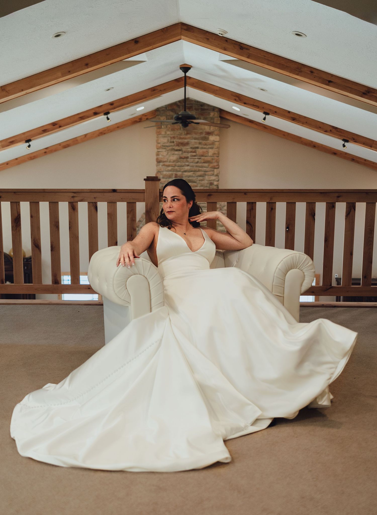 Bride in flowing white gown poses elegantly on cream couch in rustic venue with wooden beams.