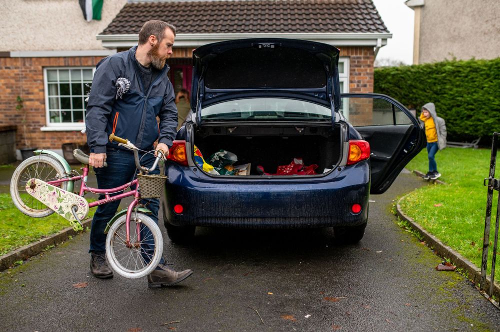Person loading a bicycle into the trunk of a dark colored car while a child stands in the background.