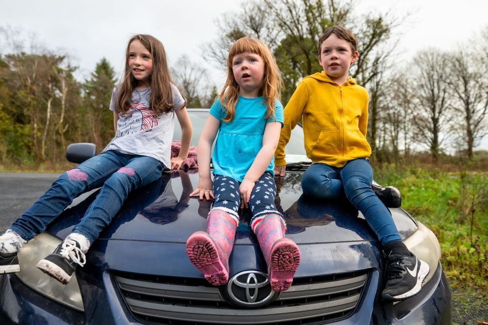 Three children sitting on hood of car wearing colorful casual clothing.