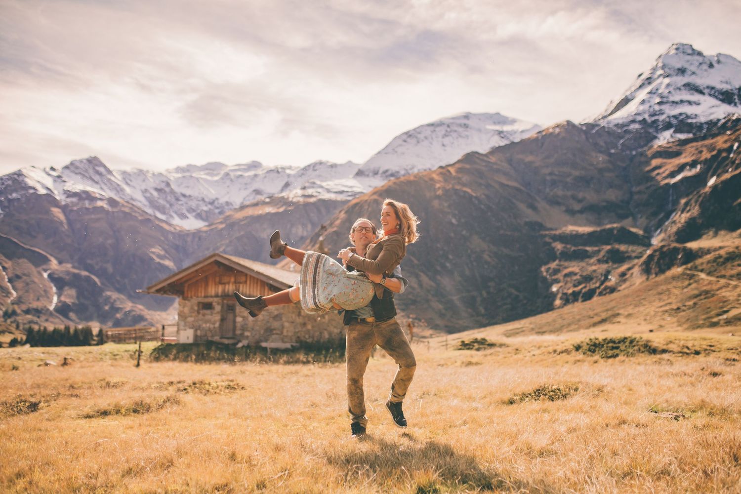 A couple embracing against a dramatic mountain landscape with snow-capped peaks in the background.