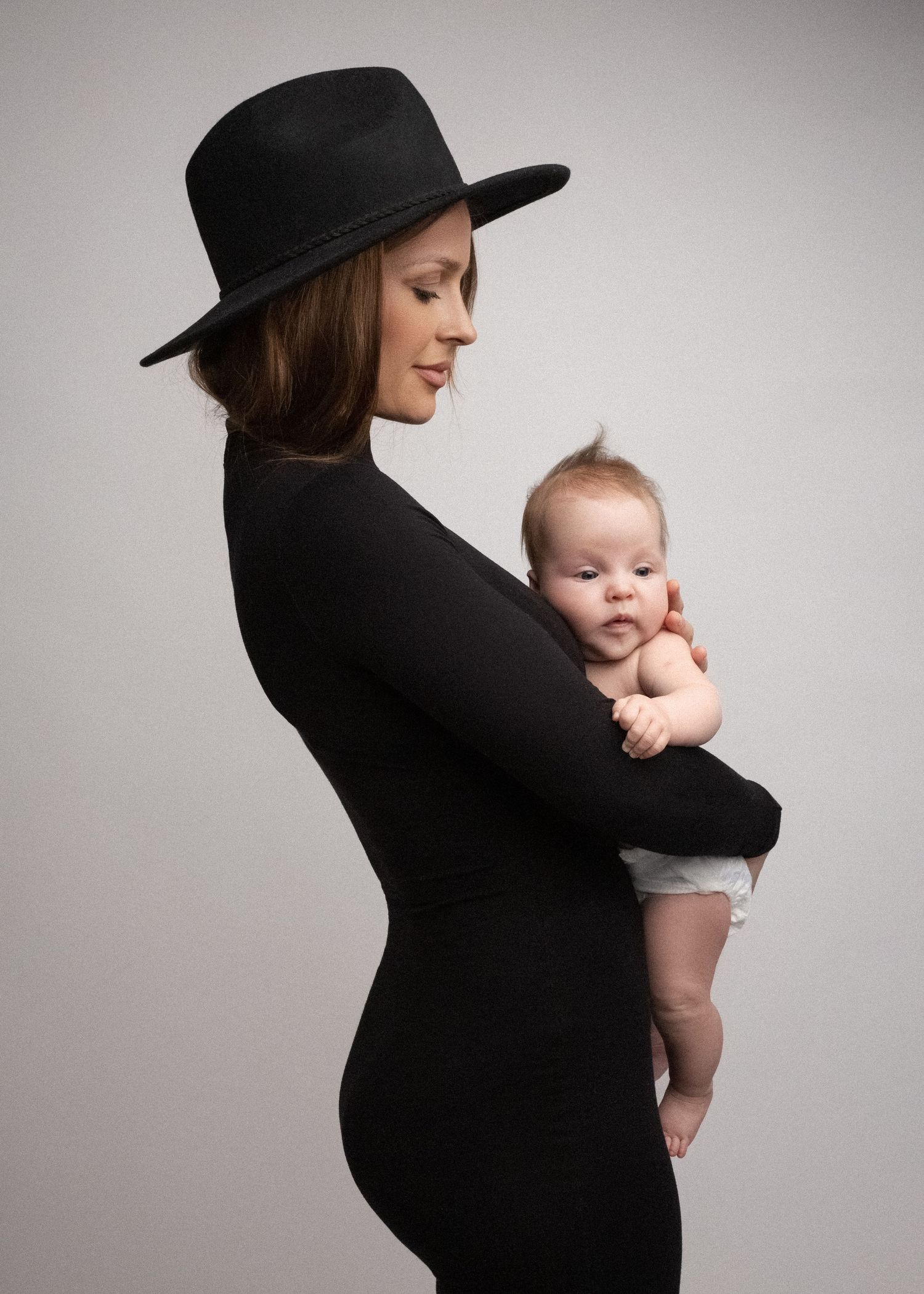 Fashion style mother and baby portrait in London by Virginija Photography. Elegant mom in a black hat holding her newborn.