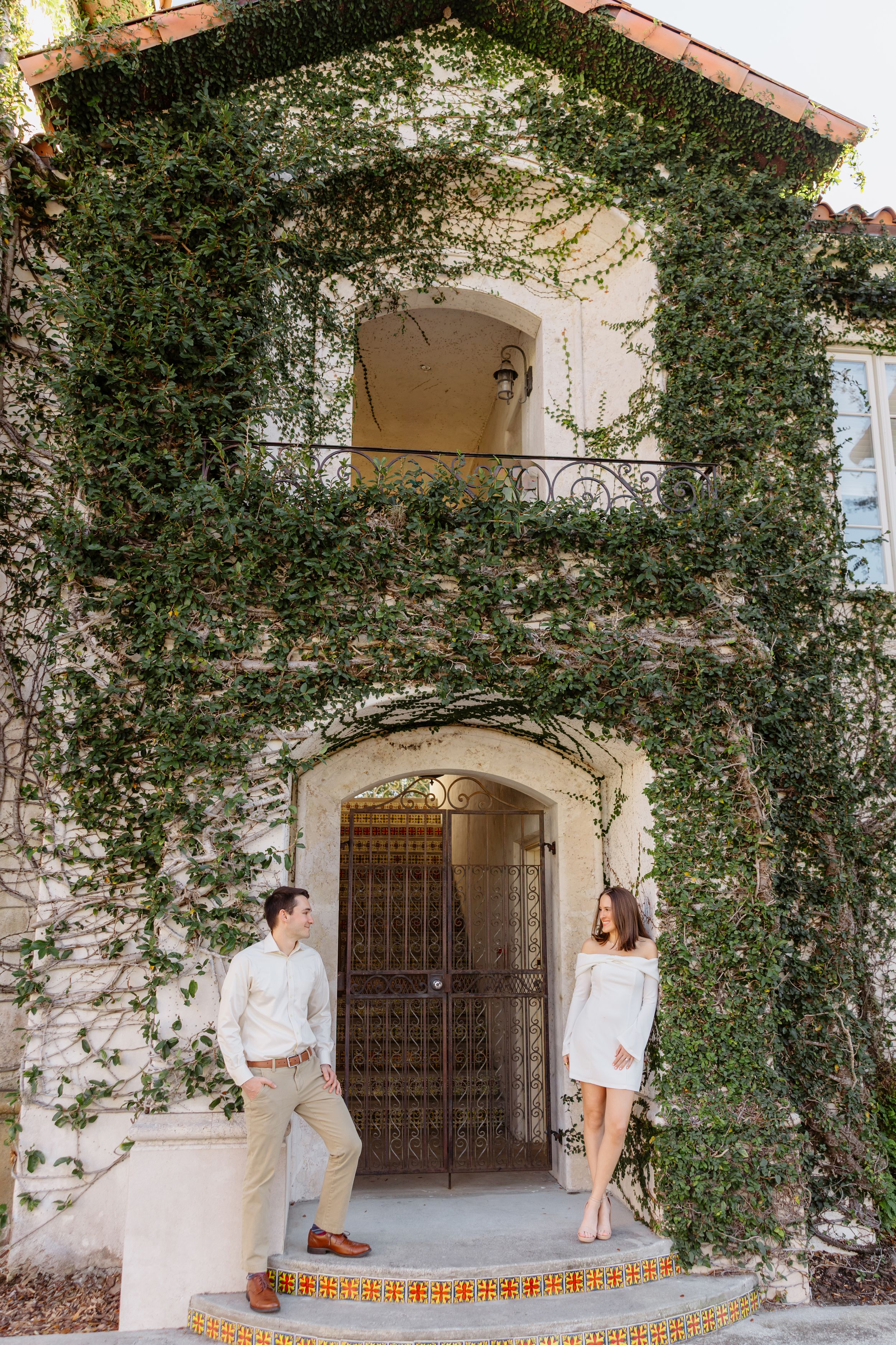 Couple taking romantic engagement photos in front of an ivy-covered Spanish style archway and courtyard entrance.