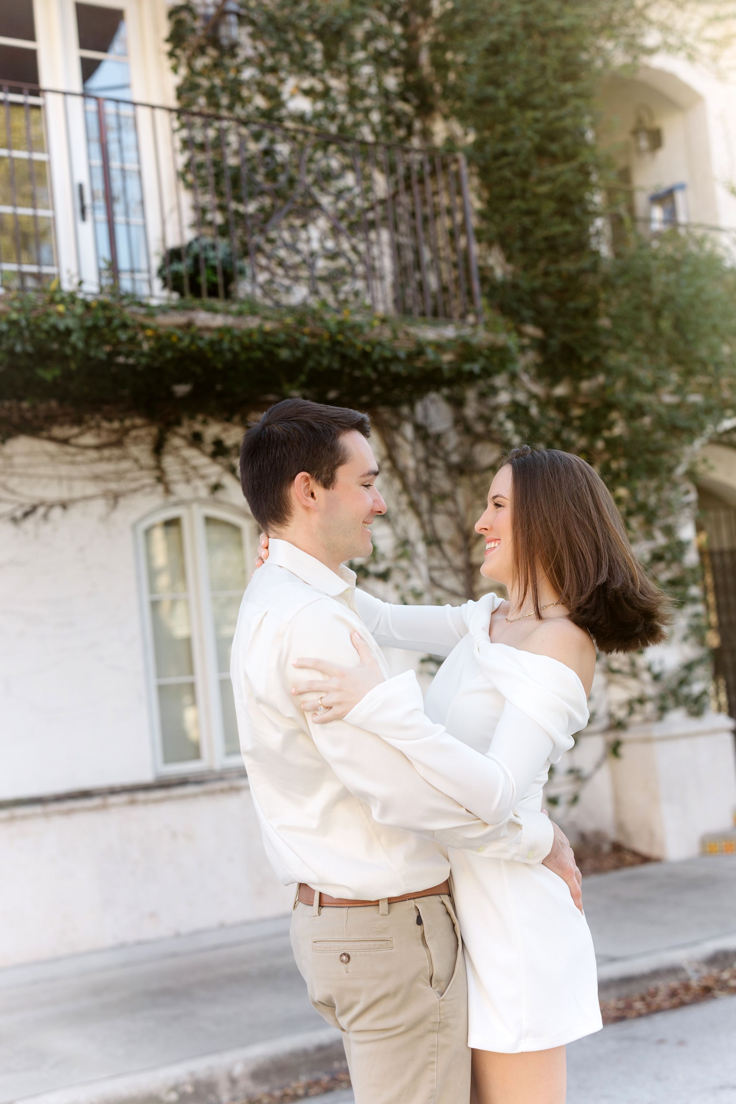 A couple in white and beige outfits dance together on a paved walkway near a building.