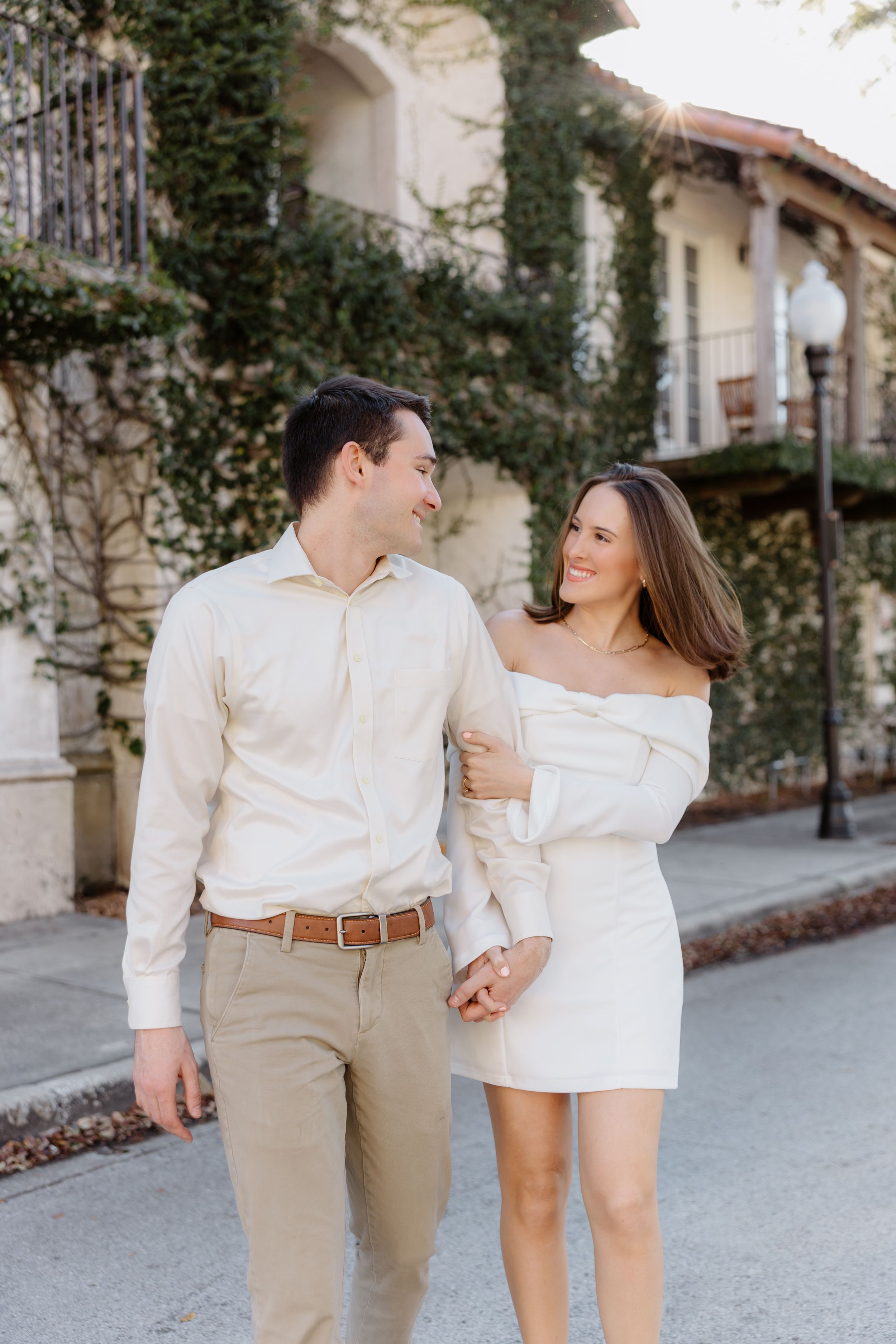 A couple in coordinating white outfits walks together on a sidewalk beside a Mediterranean-style building with climbing vines.