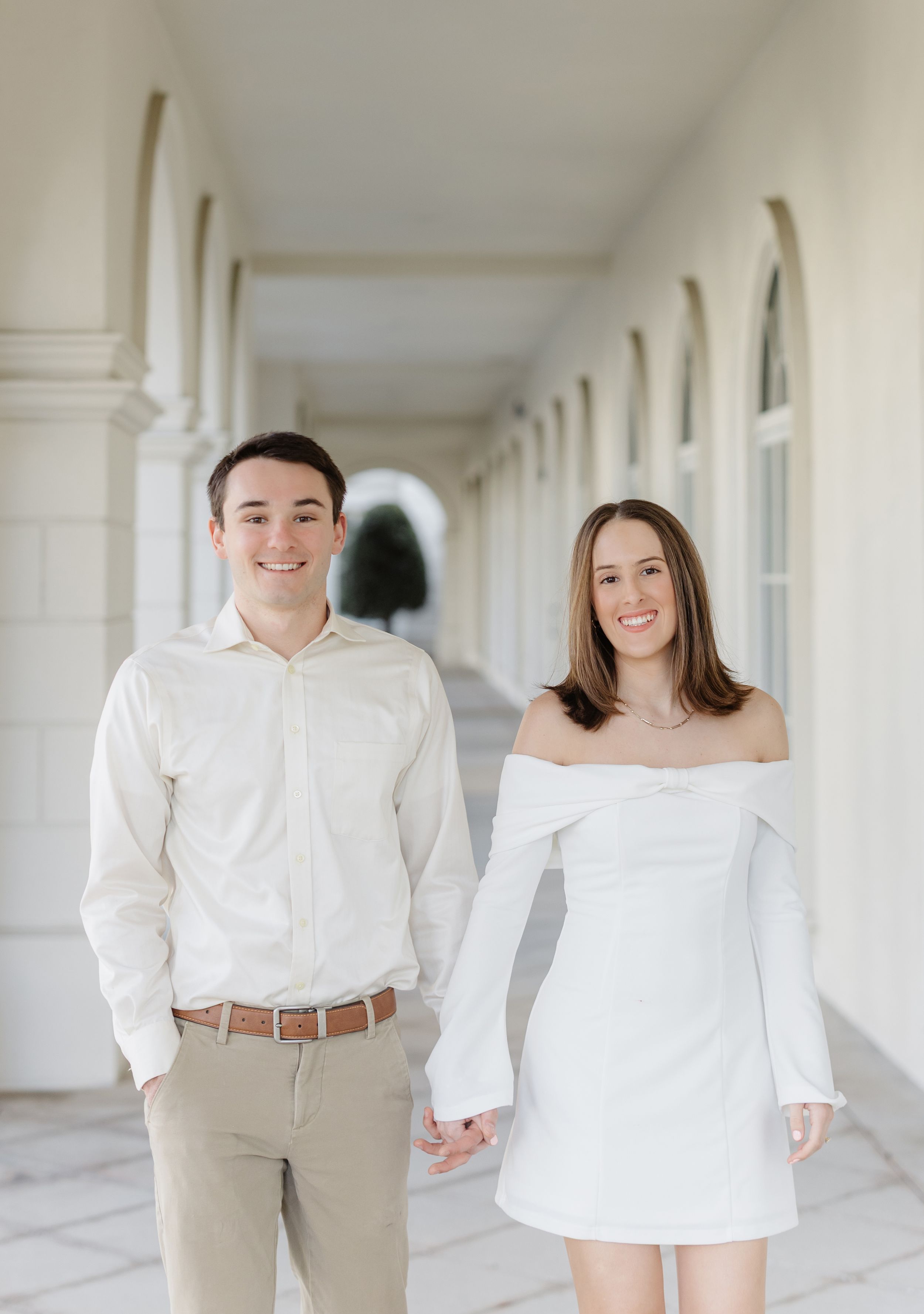 A couple in white and beige outfits poses together in a bright hallway with white columns.