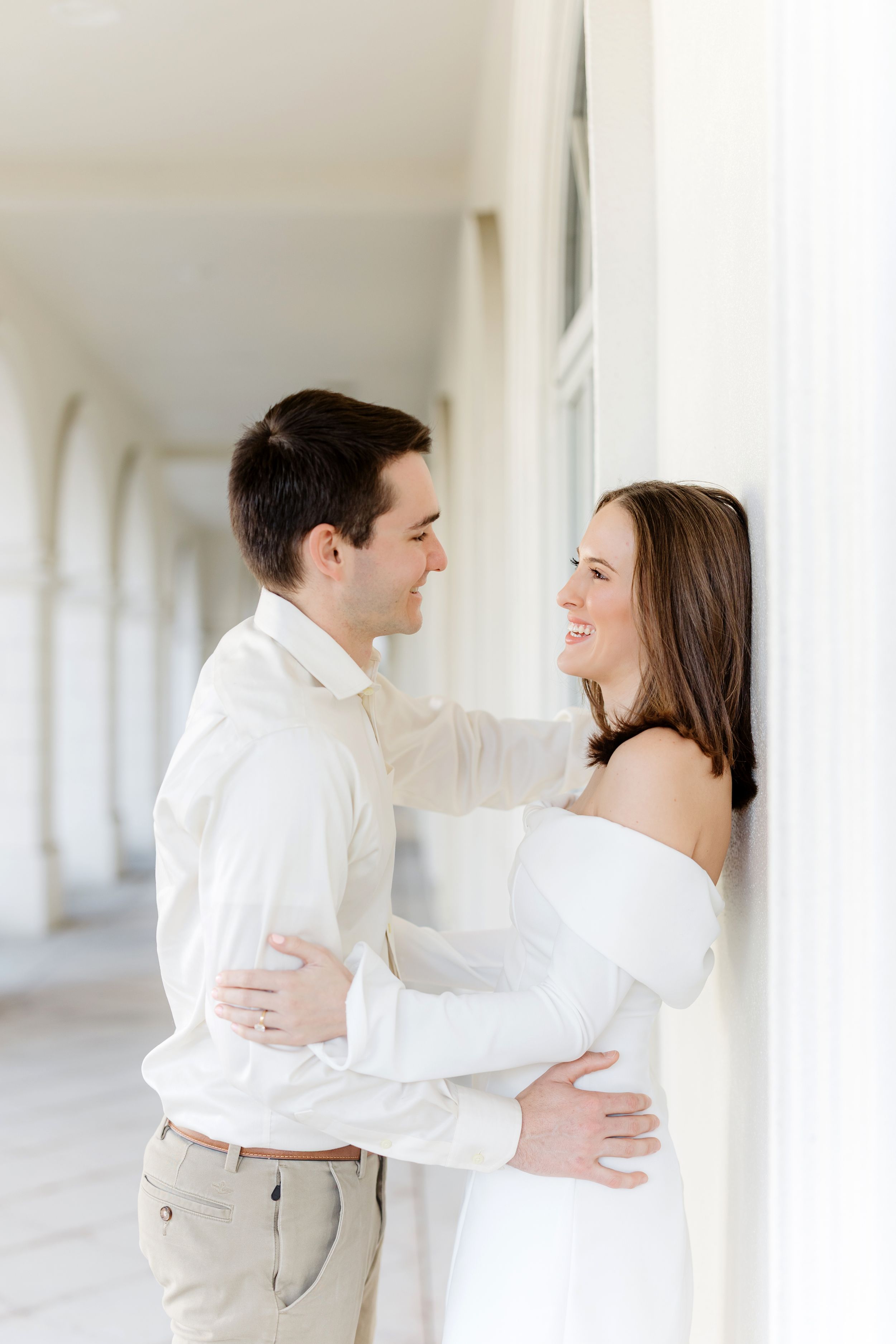 A couple in white clothing embraces and laughs together in a bright hallway with columns.