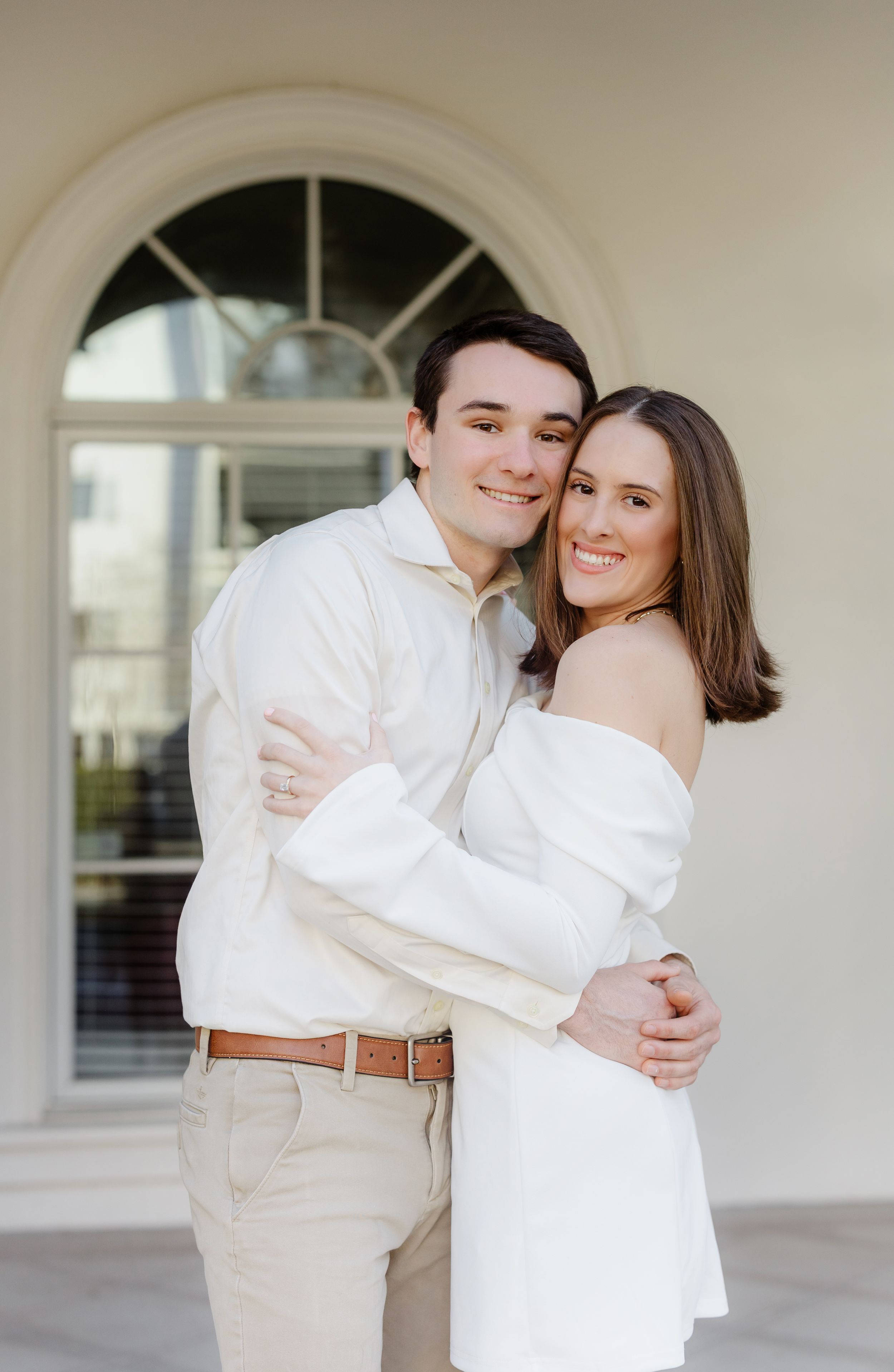 A romantic couple in white outfits embrace in front of an elegant arched colonnade.