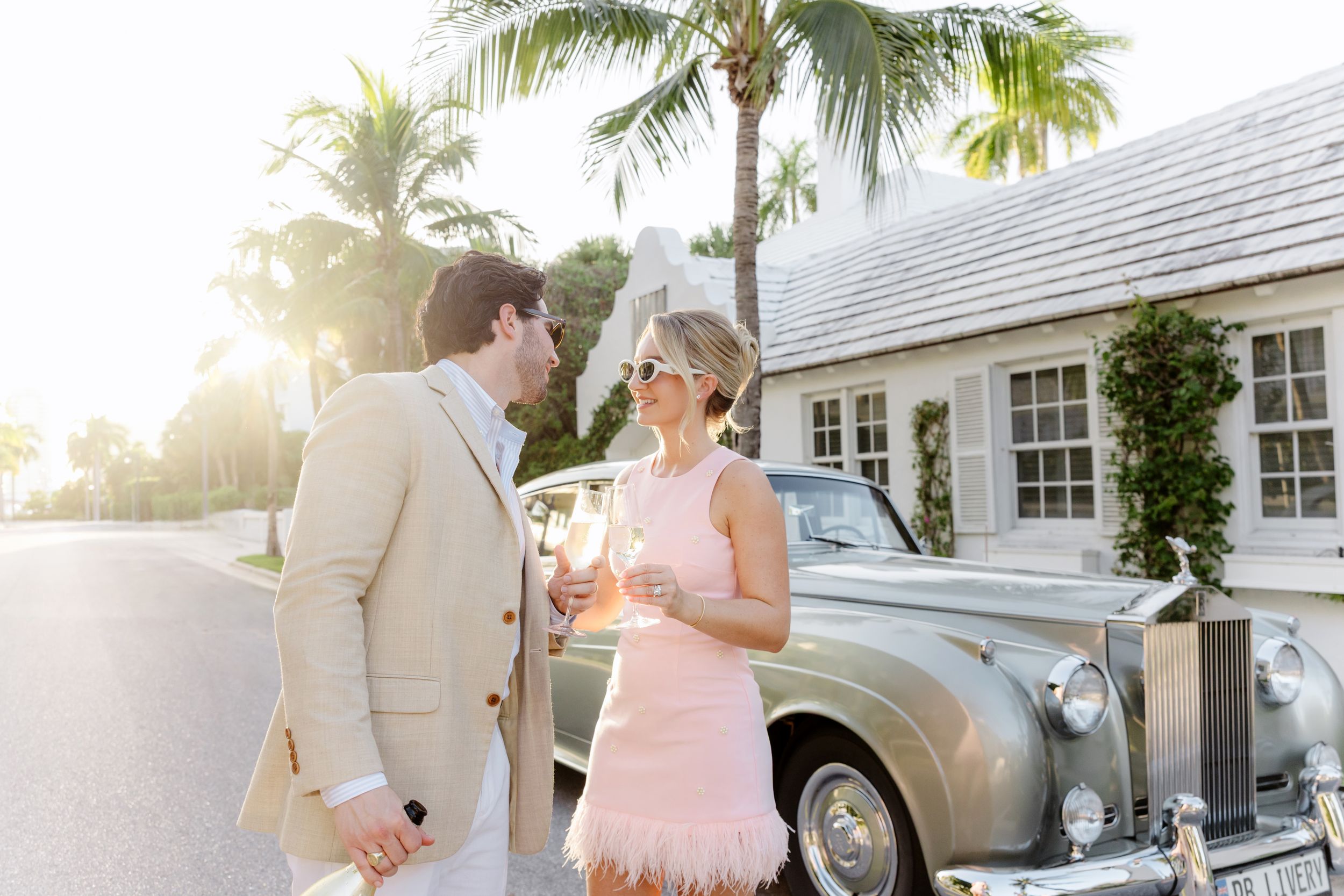 Elegant couple in formal attire holding champagne glasses beside a vintage car outside a luxury home.