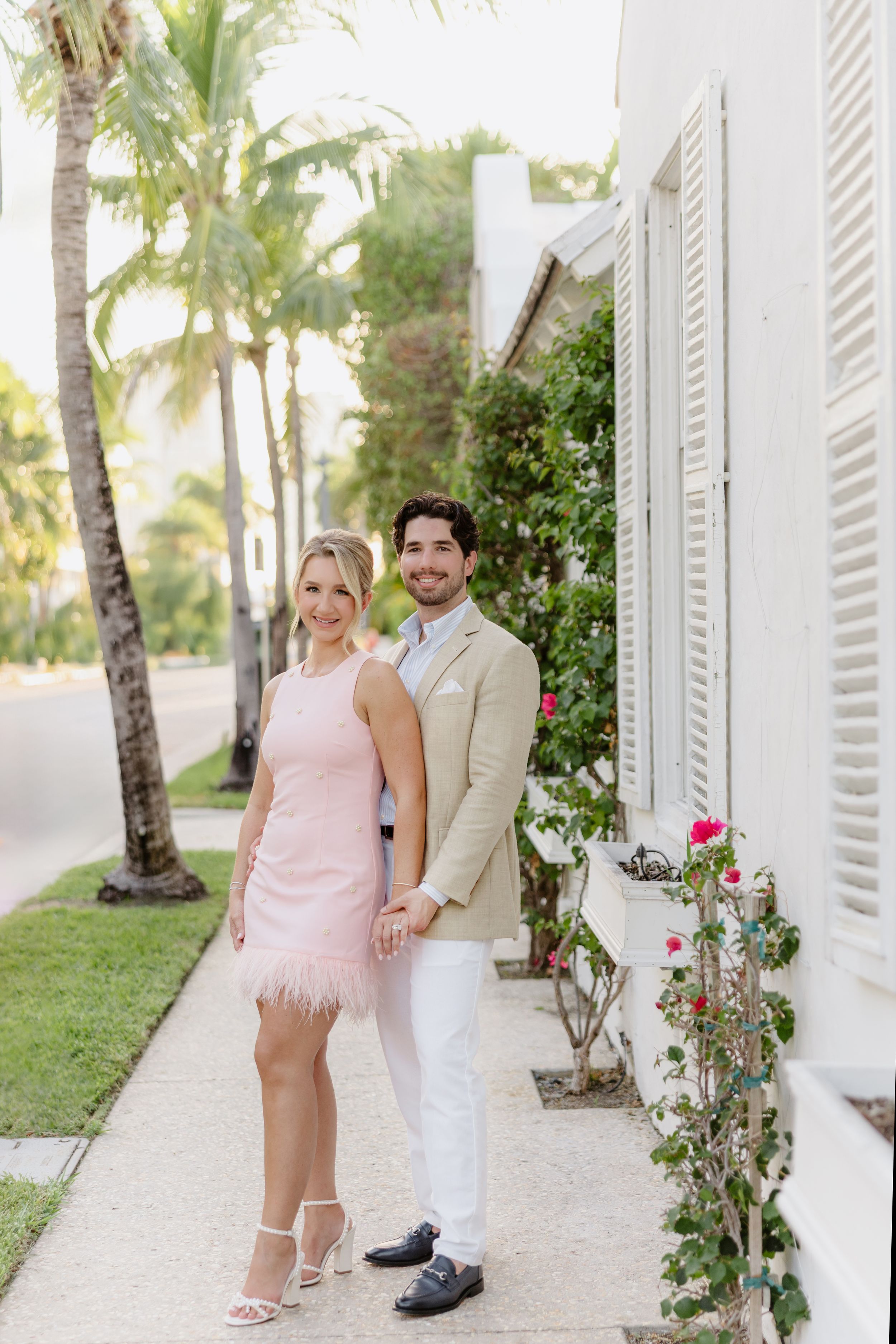 Couple posing on a sunny sidewalk, woman in pink feather mini dress, man in beige blazer and white pants, tropical setting.