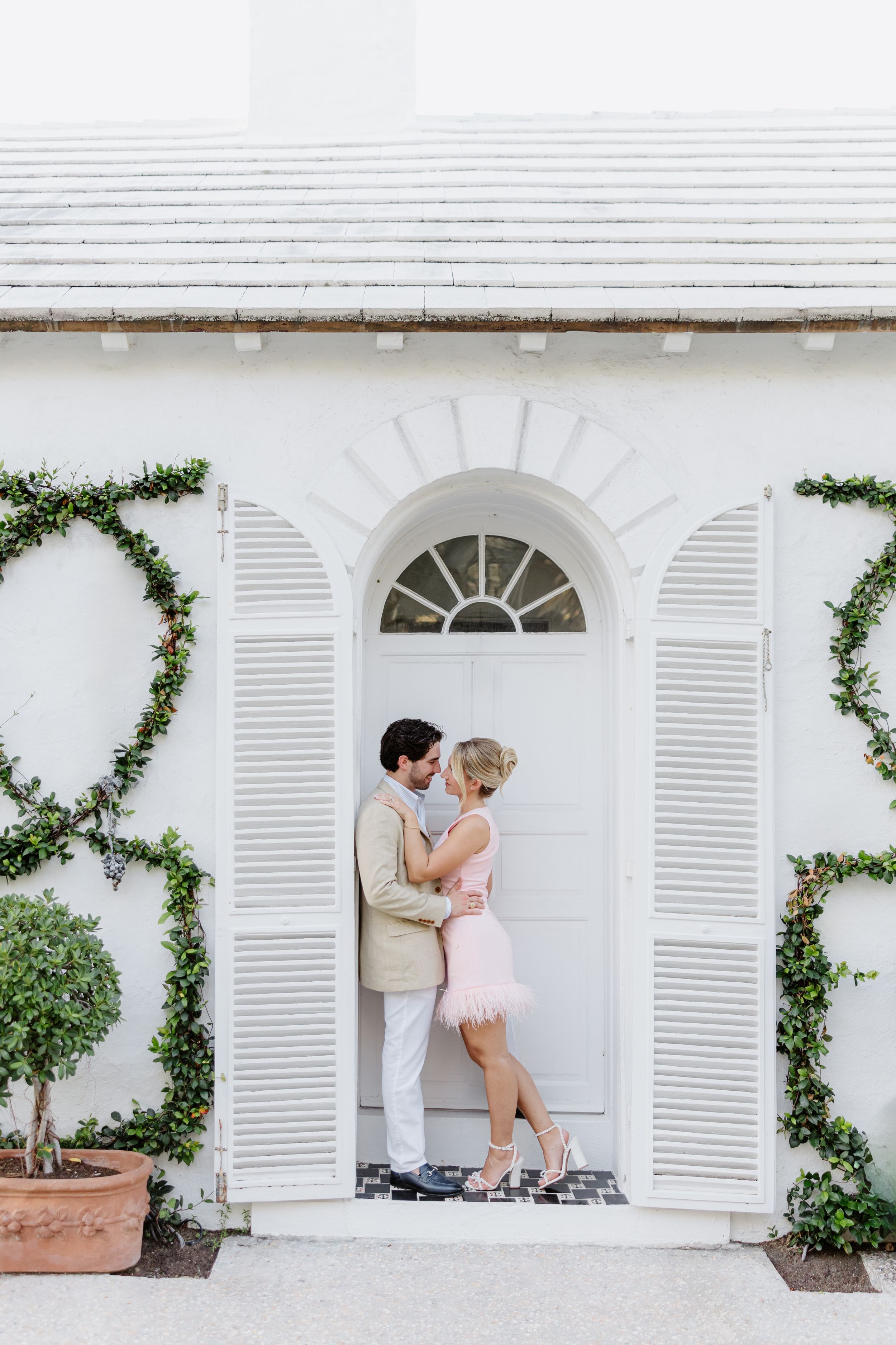 Couple sharing a romantic kiss in an elegant white arched doorway with ivy-covered walls and potted topiaries.
