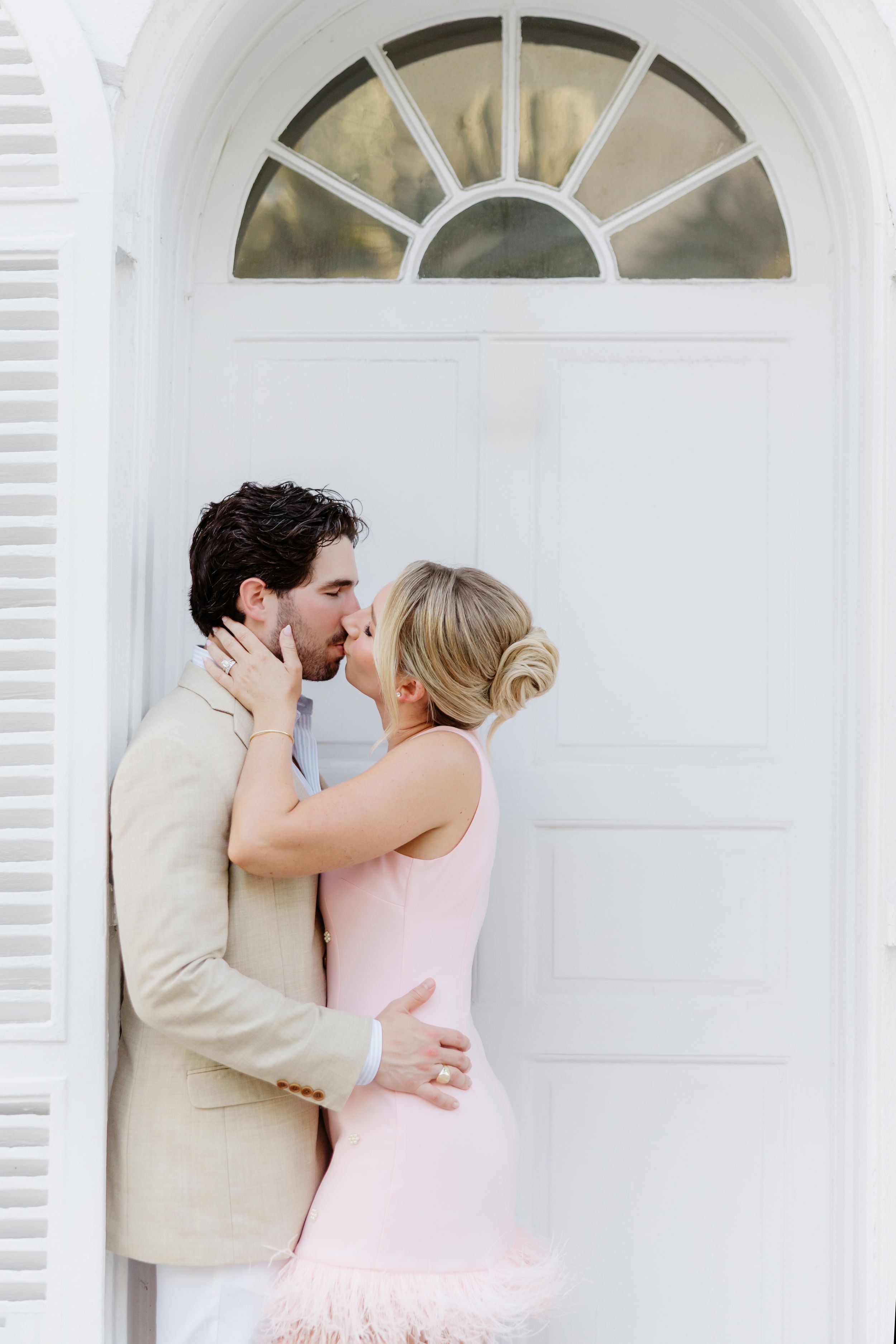 Couple sharing a romantic kiss in front of a white door, man in tan suit and woman in pink dress.