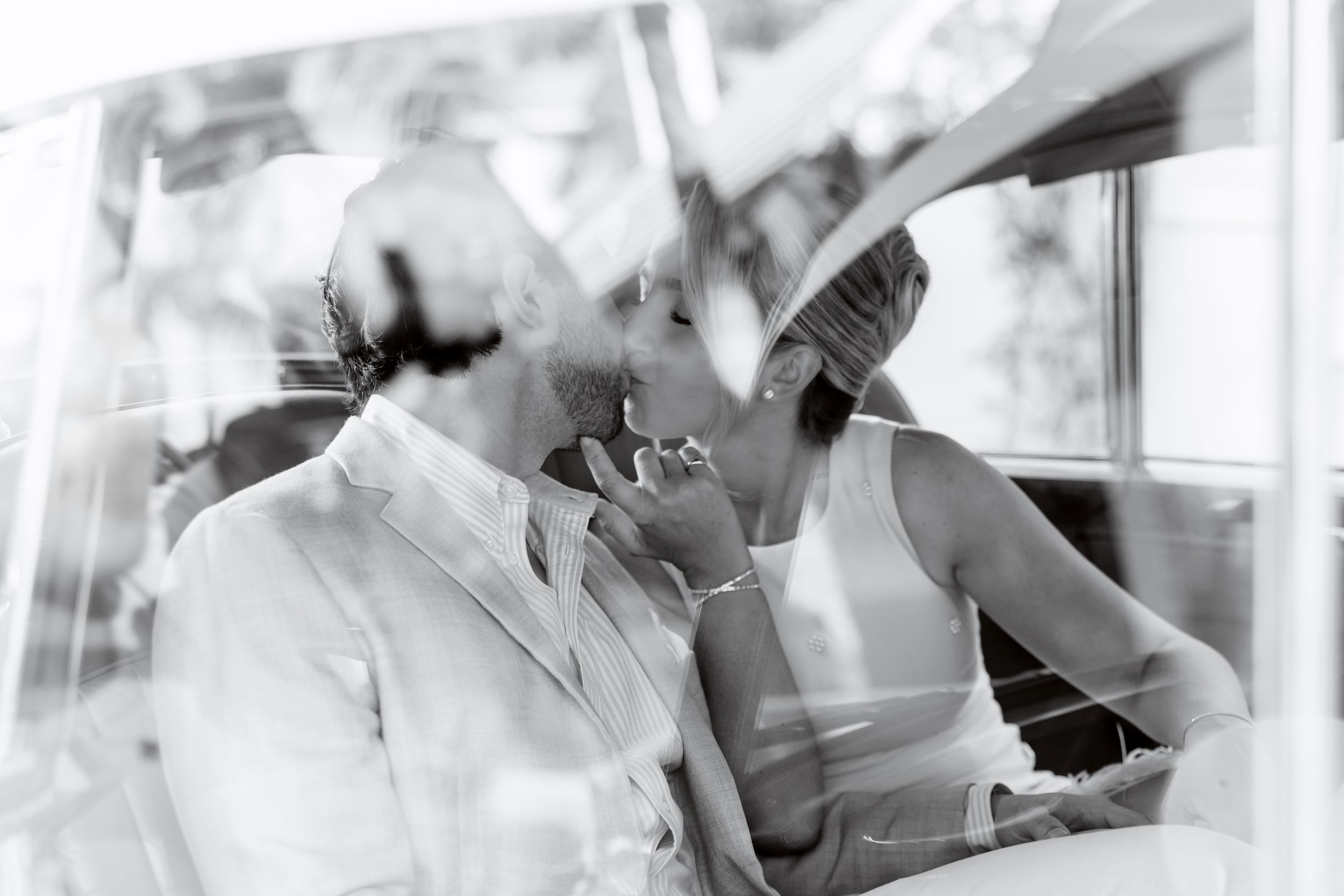Black and white photo of a couple sharing a romantic kiss inside a vintage car on their wedding day.