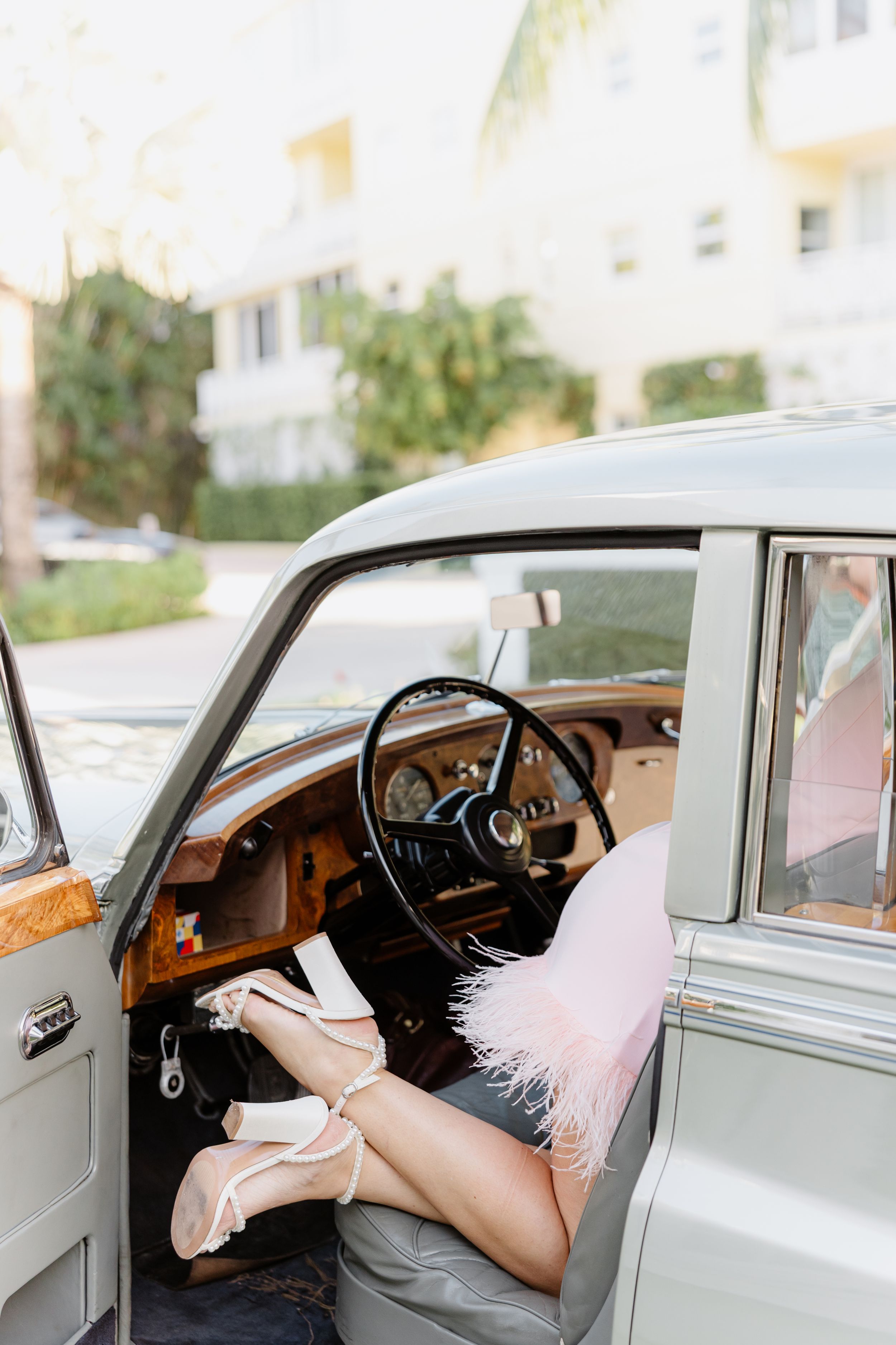 Woman in pink feather dress and heels posing in a vintage classic car with wood trim steering wheel.