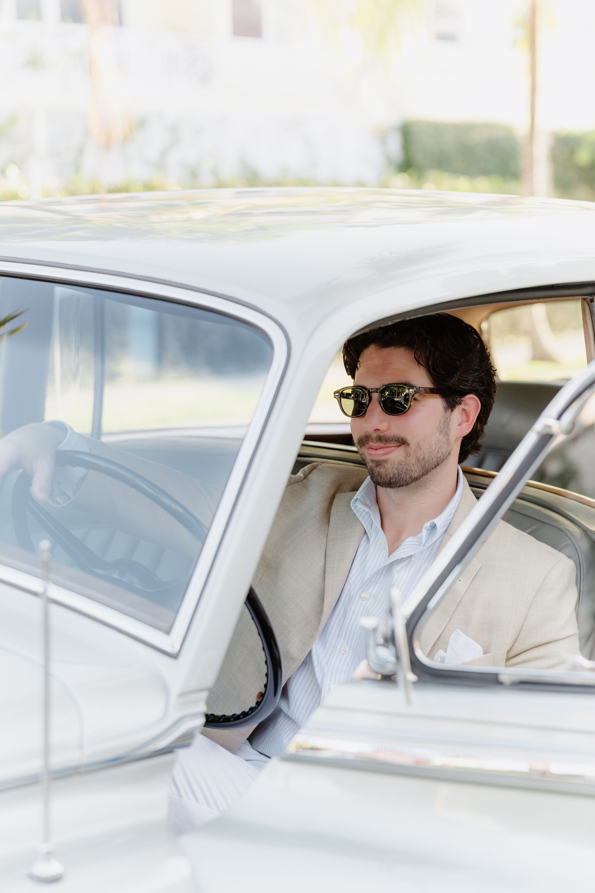 Stylish man in beige blazer and sunglasses sitting in a classic white vintage car, smiling confidently.