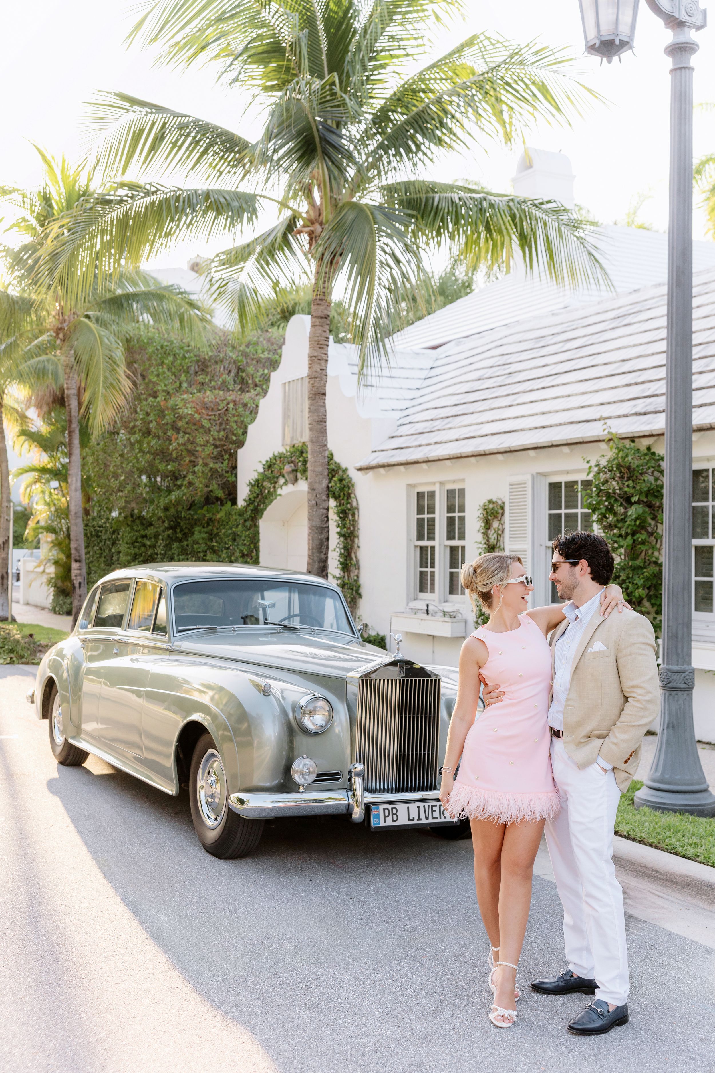Couple posing beside a vintage Rolls-Royce on a palm-lined street, woman in pink dress, man in beige suit.