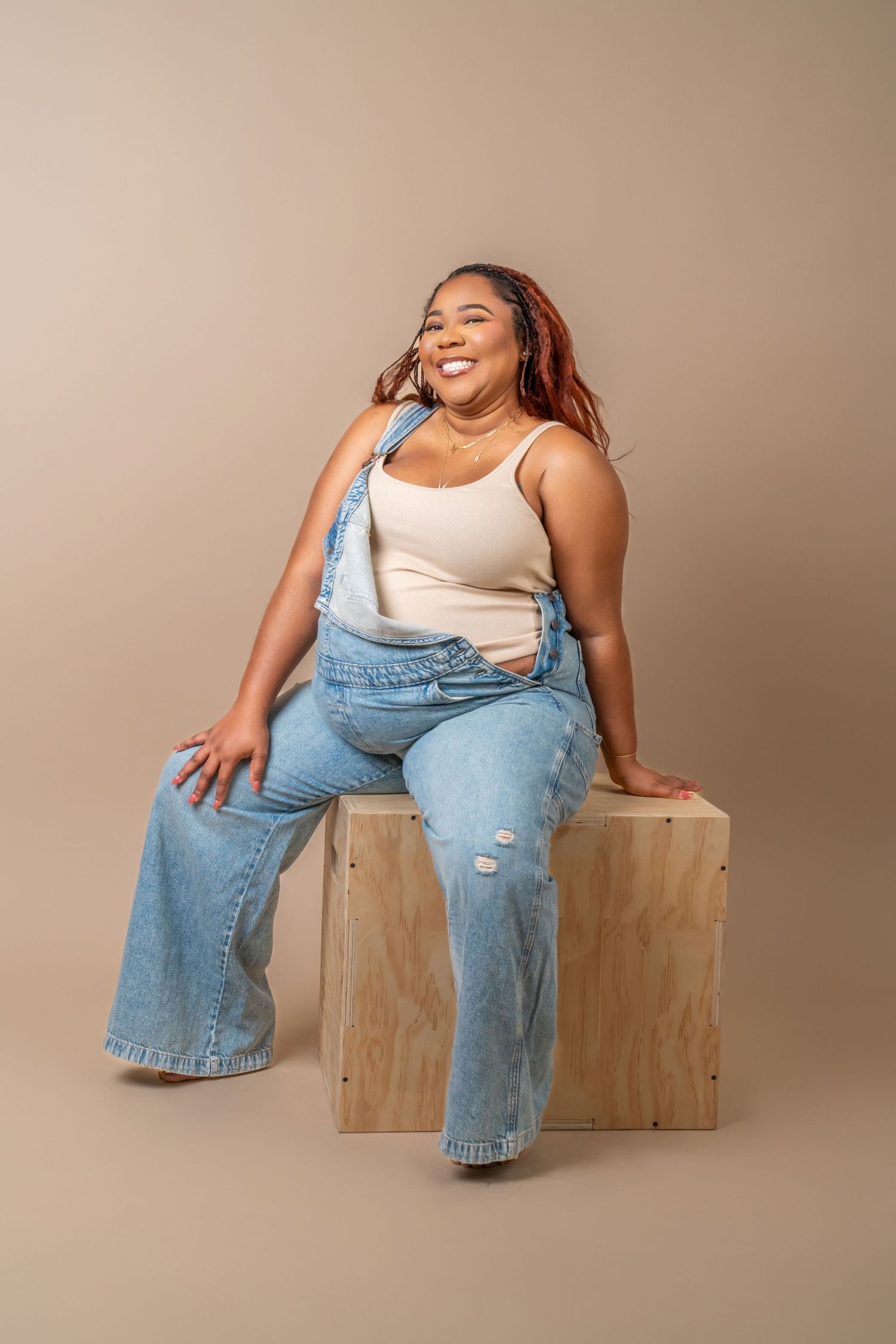 Smiling plus-size woman in denim overalls and cream tank top sitting on wooden box against beige background.