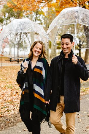 A couple walking together on a rainy autumn day, sharing clear umbrellas and wearing coordinated outfits. Philadelphia engagement photos.