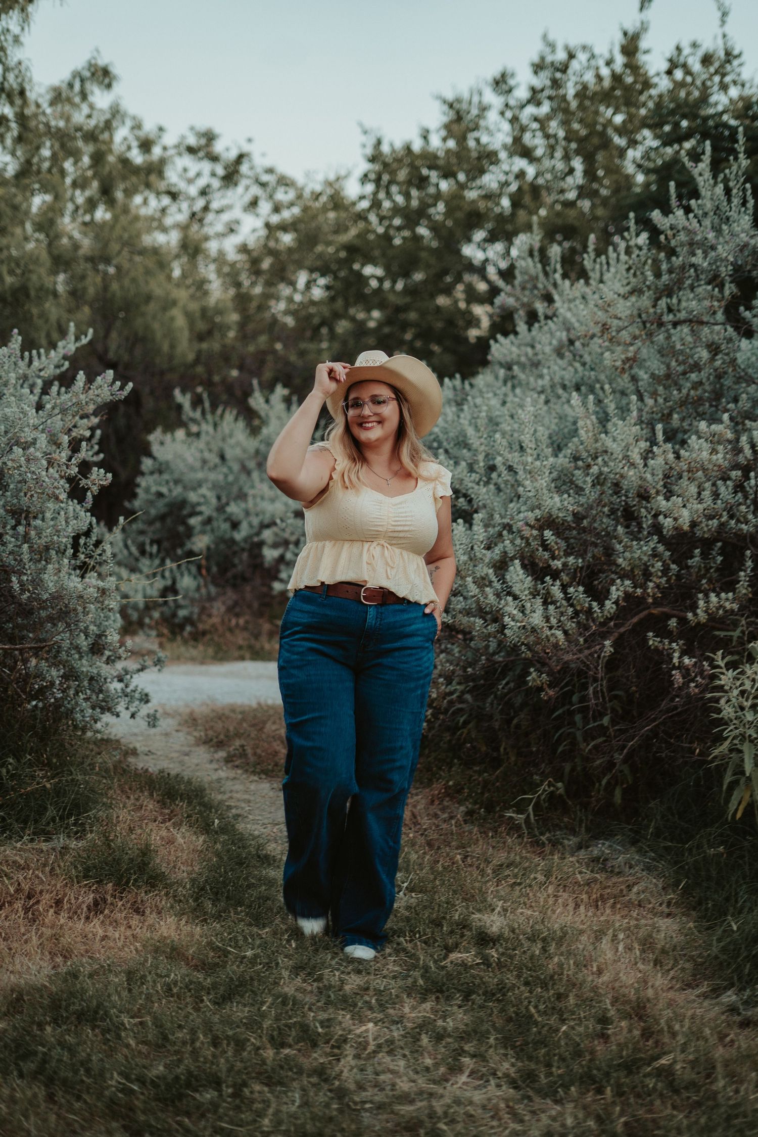 Woman in yellow top, wide-leg jeans, and straw hat walking on a nature path surrounded by lush greenery.