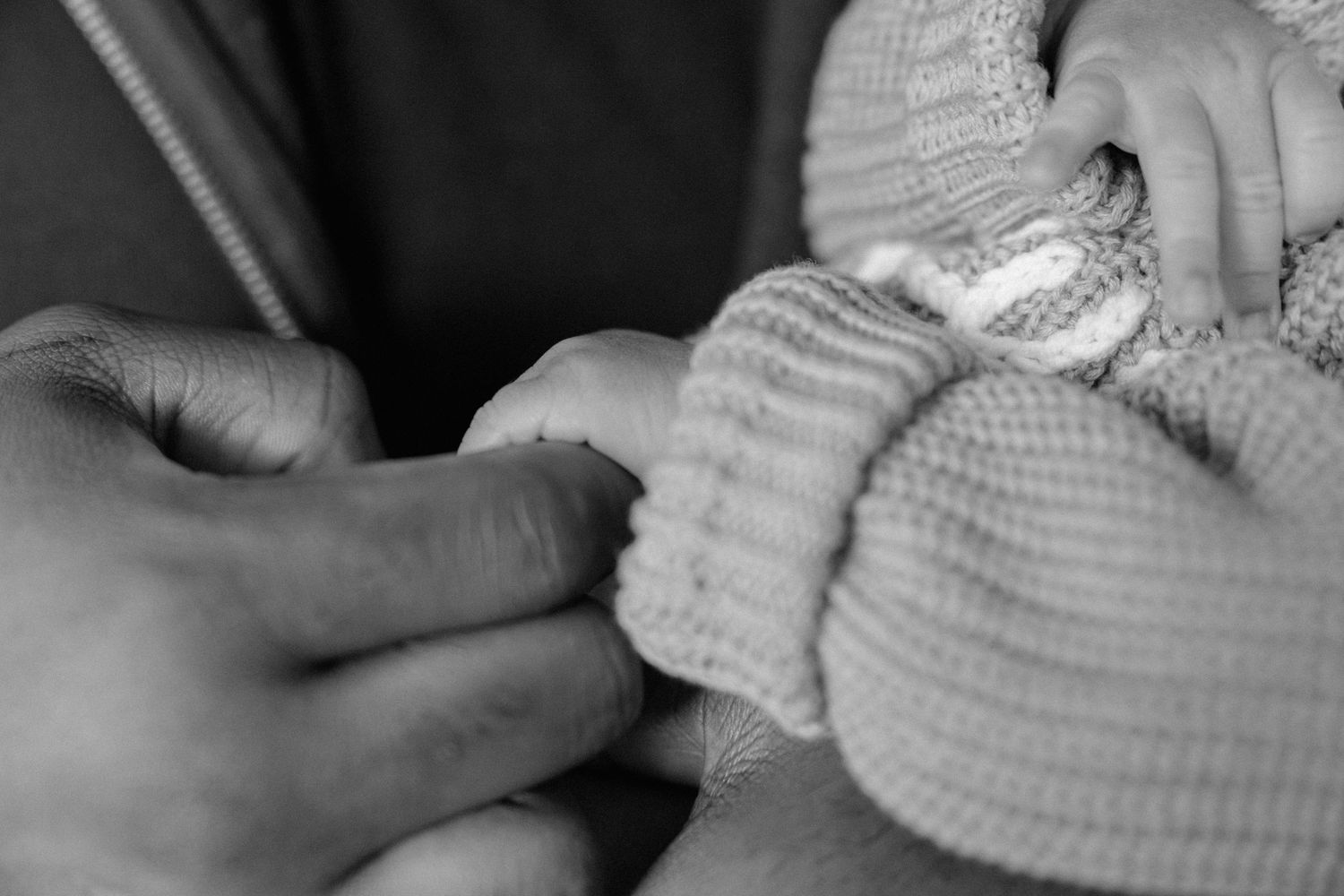 Black and white close-up of an adult hand gently holding a newborn baby's tiny hand in a knit outfit.
