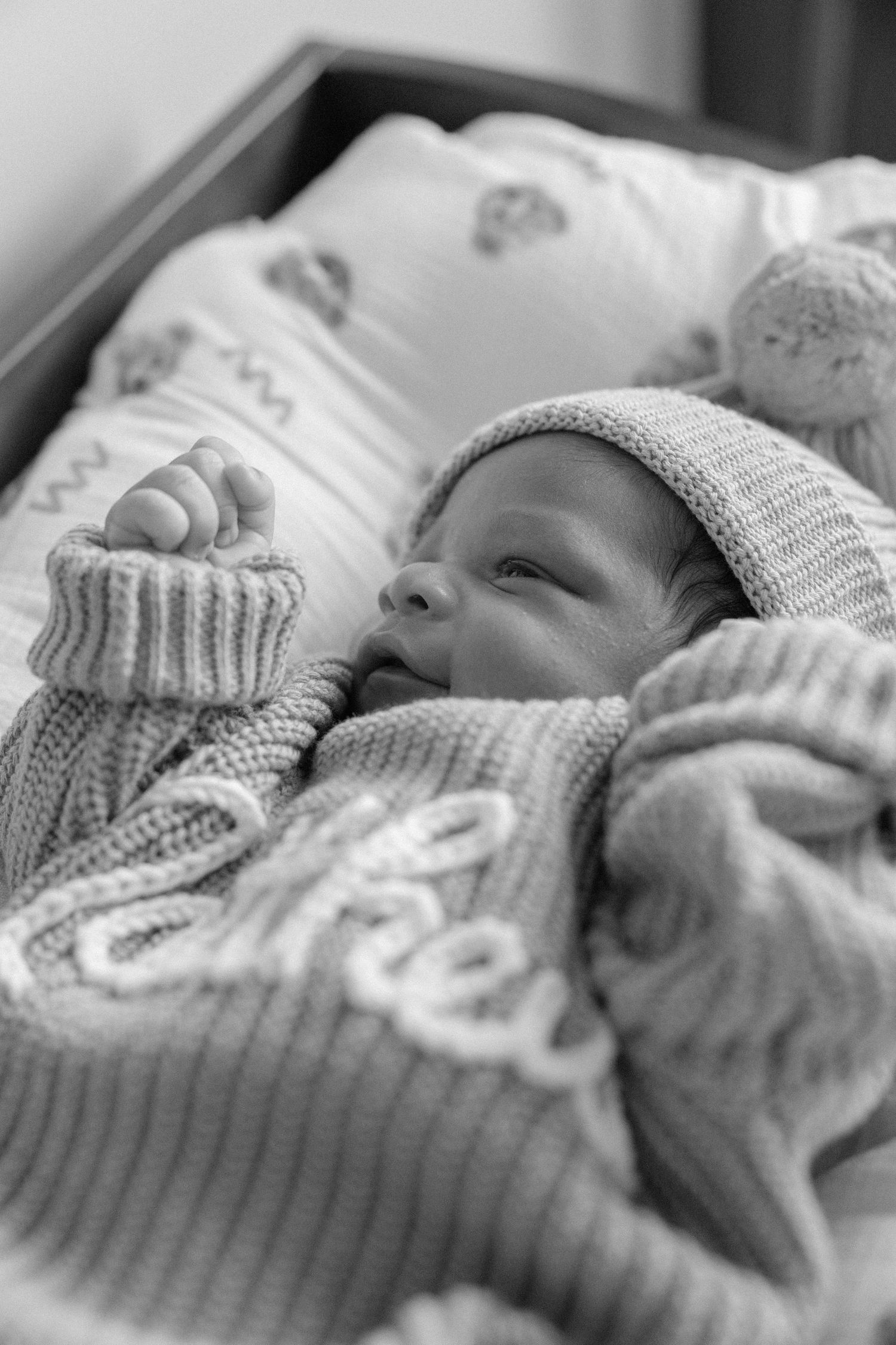 Black and white photo of a peaceful newborn baby in a knit hat and sweater lying in a crib.