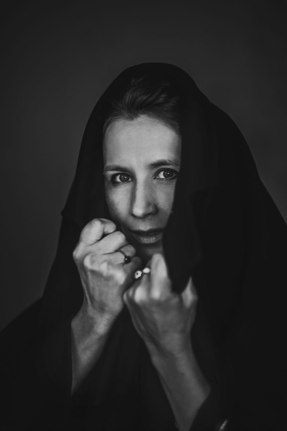 Dramatic black and white portrait of figure in dark fabric with hands raised near face. Bellevue studio, WA