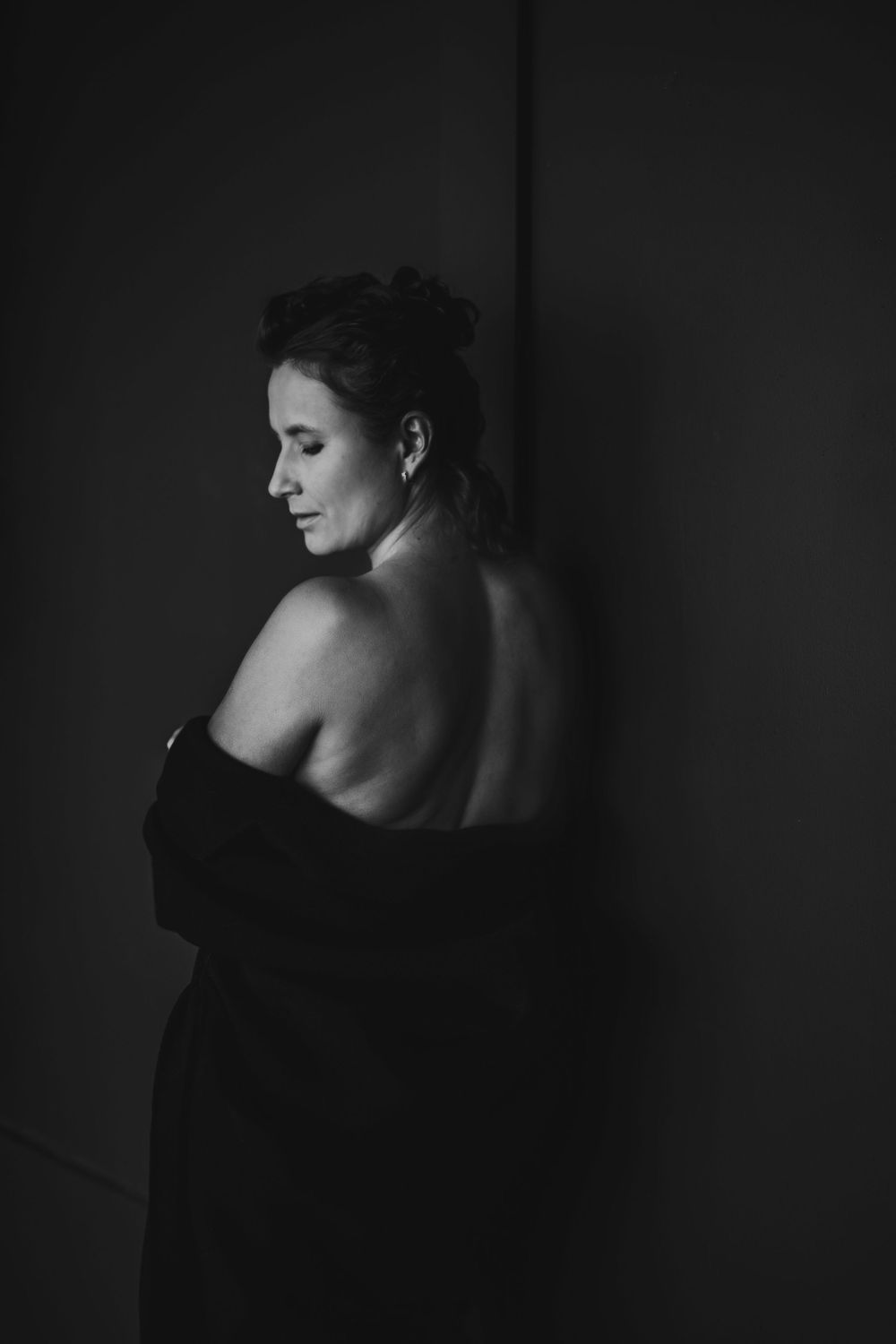 Elegant back-facing portrait showing bare shoulders in dark fabric against shadowy background. Bellevue studio, WA