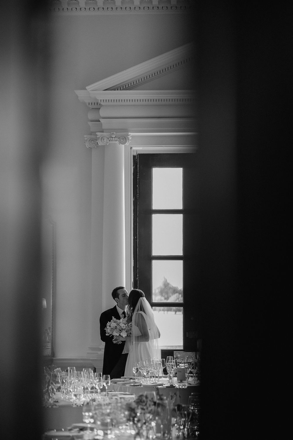 A silhouetted wedding couple embracing by a tall window with wine glasses in foreground.