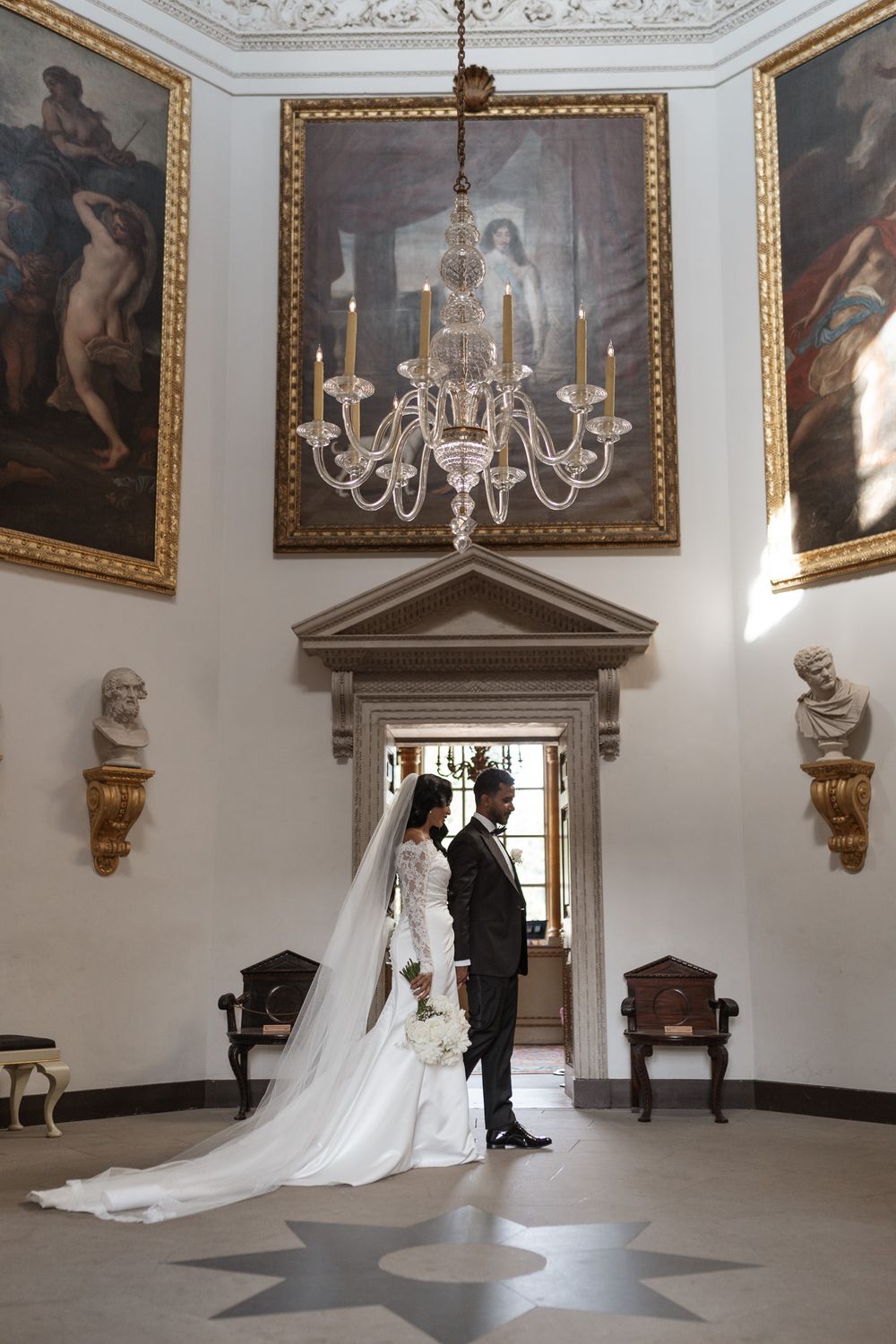 Wedding couple silhouetted in ornate room with chandelier and classical artwork.