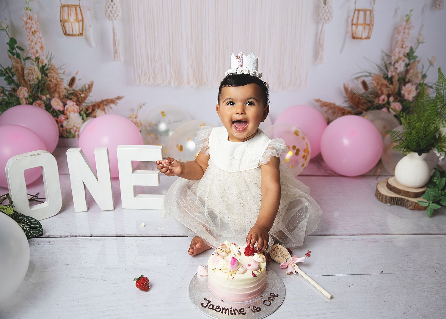 A baby in white dress with crown celebrates first birthday with cake smash setup featuring pink balloons and floral decor in Bexley