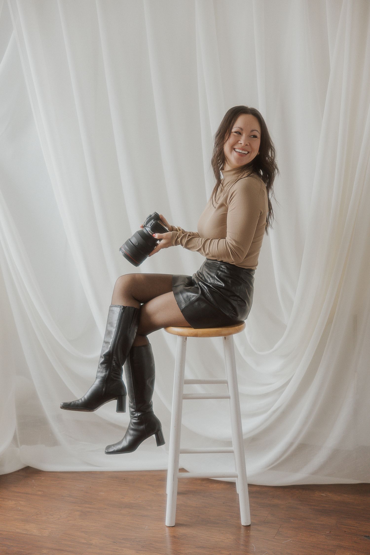 Professional photographer poses on a stool with camera against white backdrop wearing boots and sweater.
